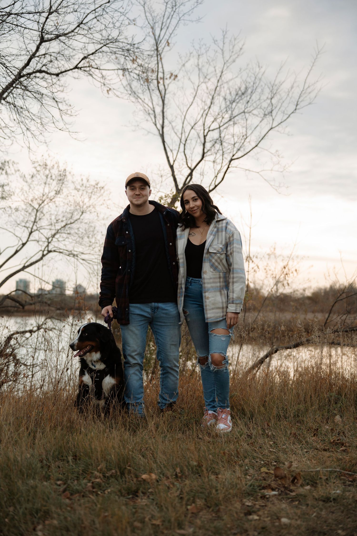 Couple standing with their dog near a lake surrounded by bare trees and tall grass during a cloudy autumn day.