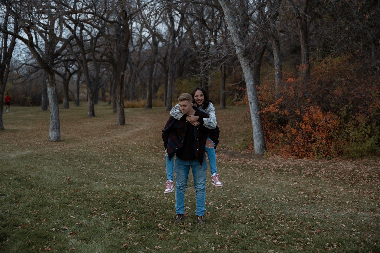 Two people posing for a photo in a park with bare trees and autumn foliage in the background.
