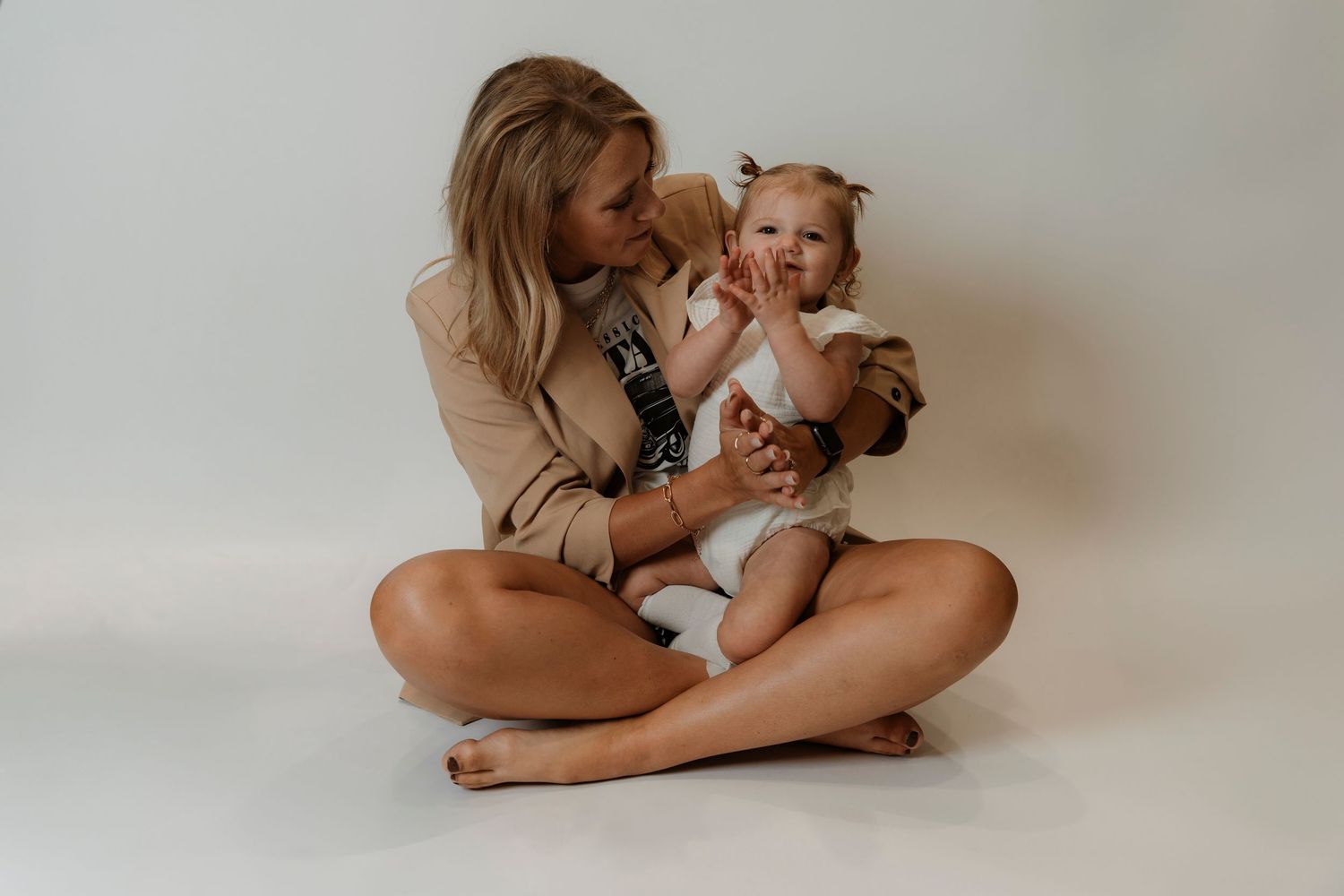 A young woman sits cross-legged on a white floor, lovingly kissing a baby girl in a white onesie.