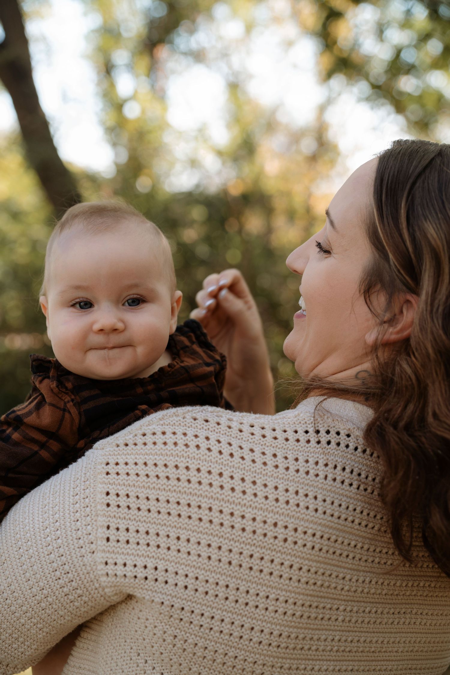 A mother in a cream knit sweater holds her smiling baby outdoors among autumn trees.