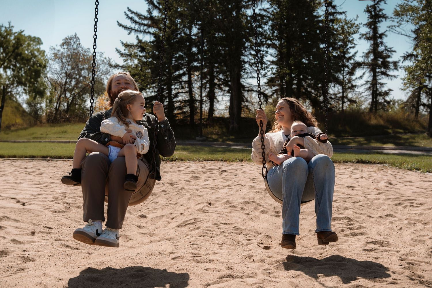 Two women sitting on swings at a sunny playground, smiling and chatting while holding babies outdoors.