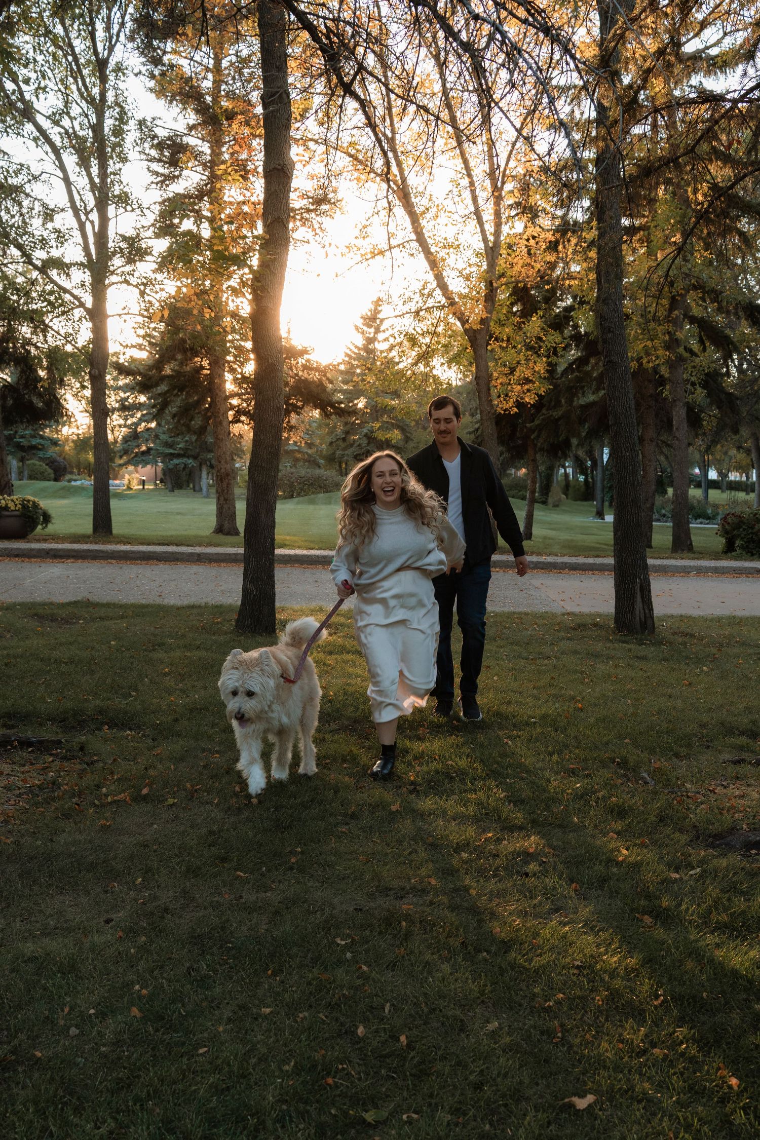 Couple walking a fluffy white dog in a sunlit park during golden hour, surrounded by tall trees and green grass.
