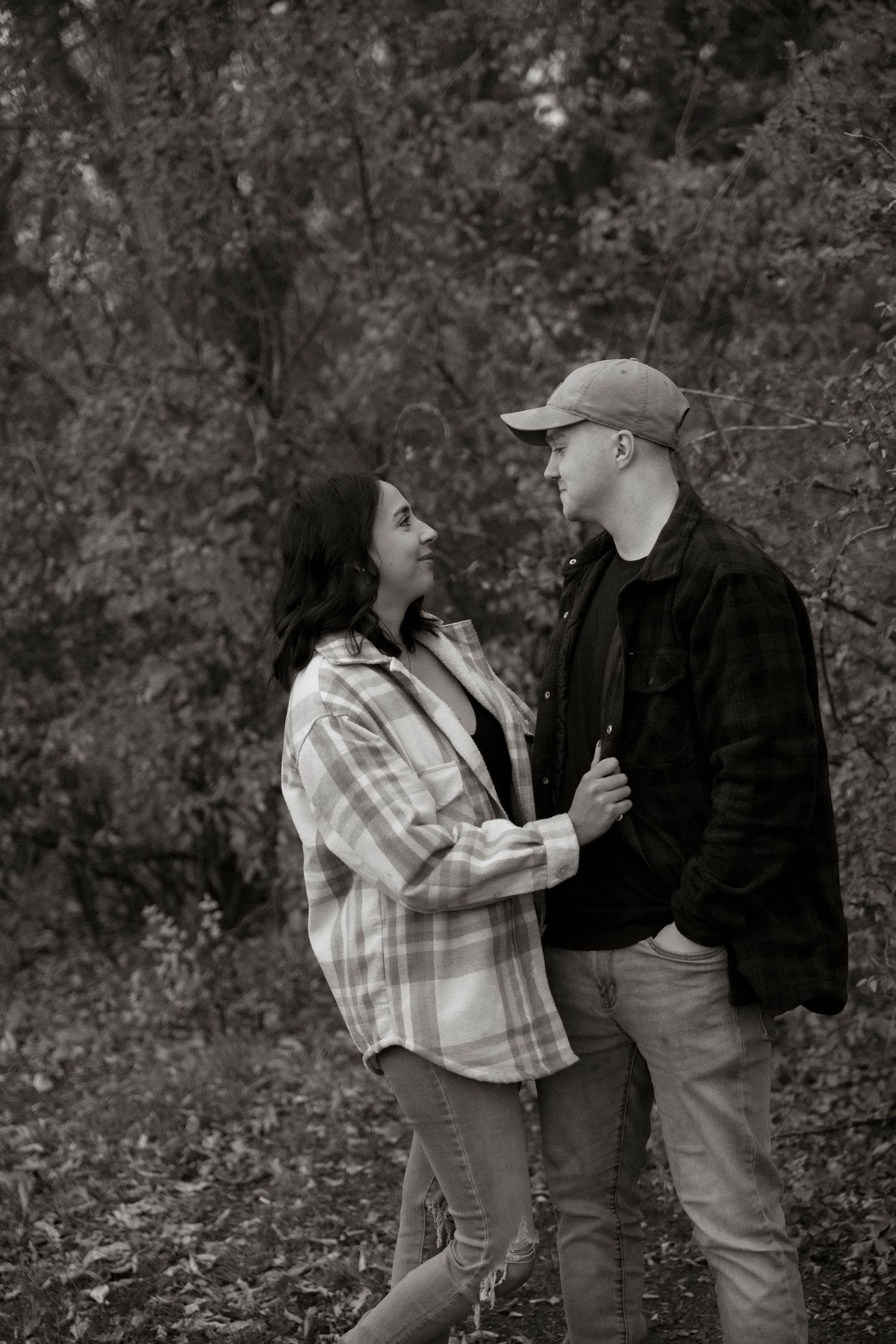 Black and white photo of a couple holding hands and gazing at each other in a wooded outdoor setting.