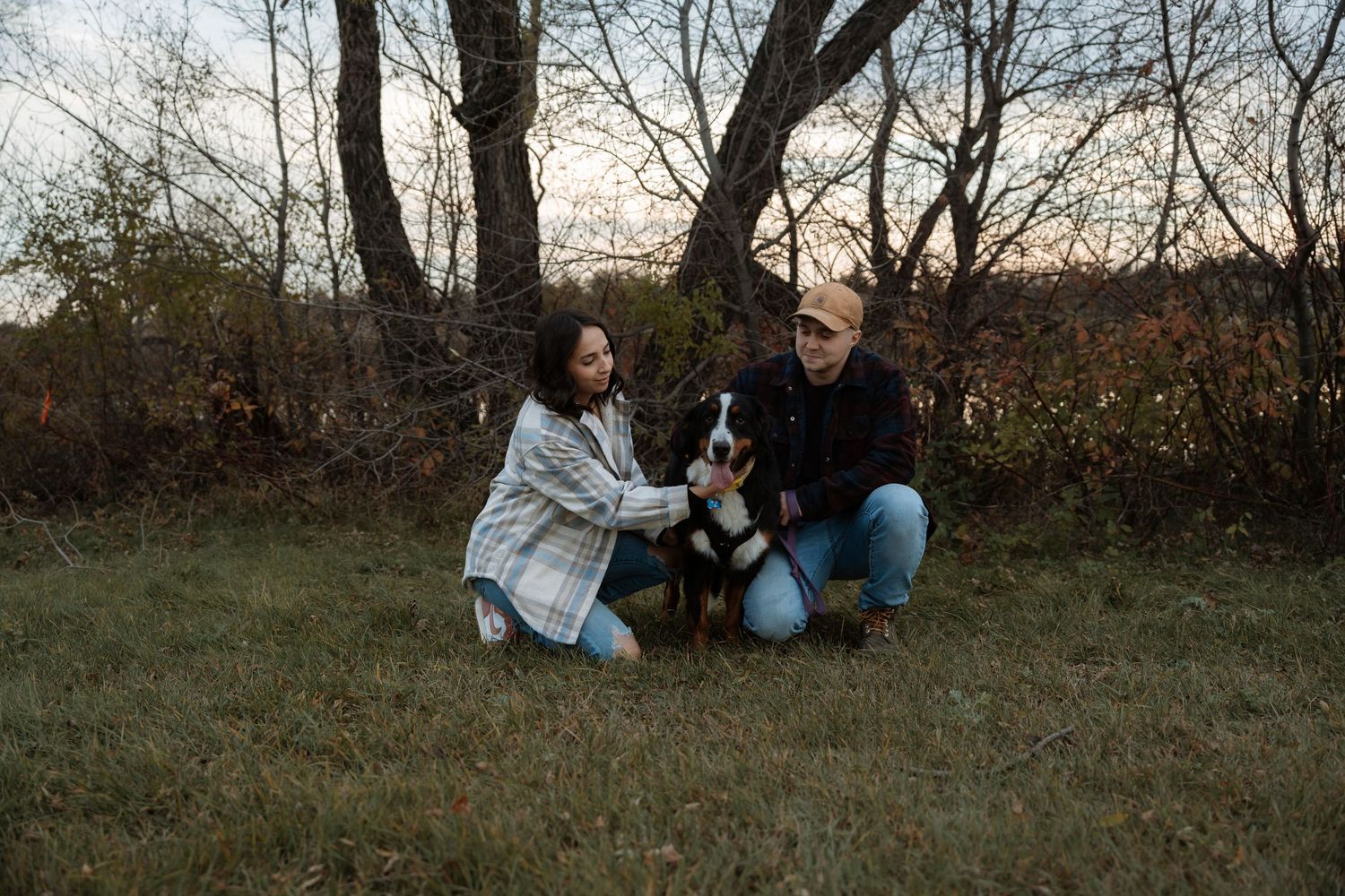 Couple kneeling with a Bernese Mountain Dog in an autumn field surrounded by bare trees at dusk.