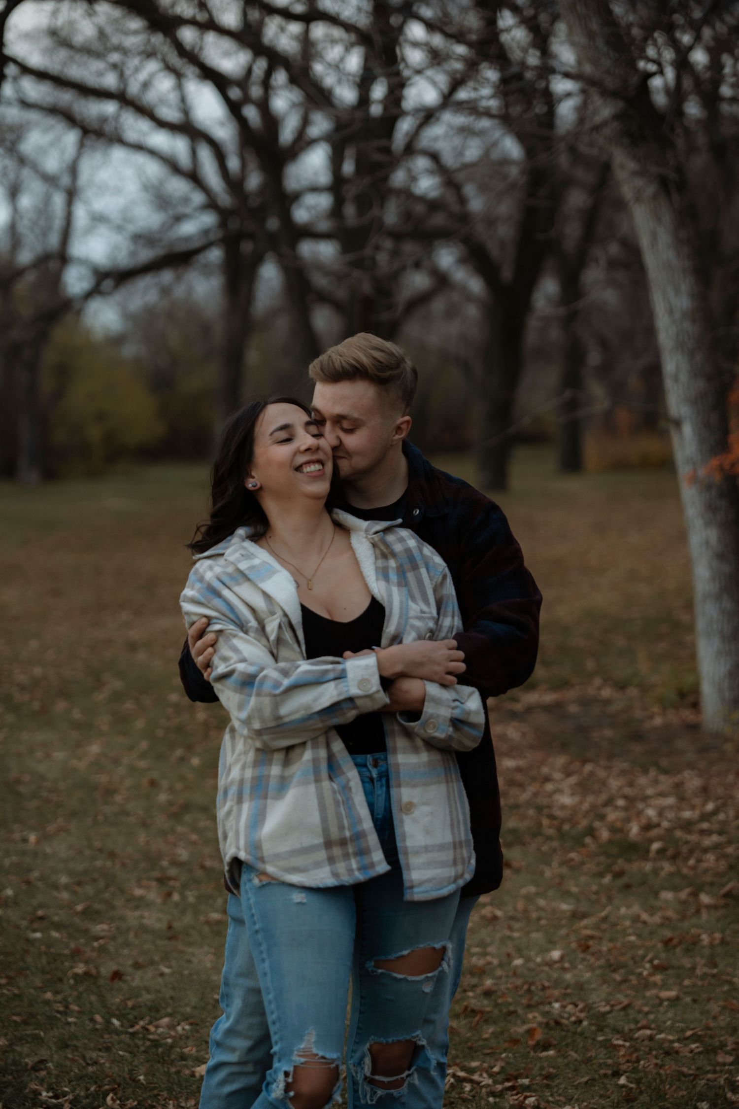 Couple embracing in autumn park, man kissing woman's cheek, wearing casual flannel and jeans among bare trees.