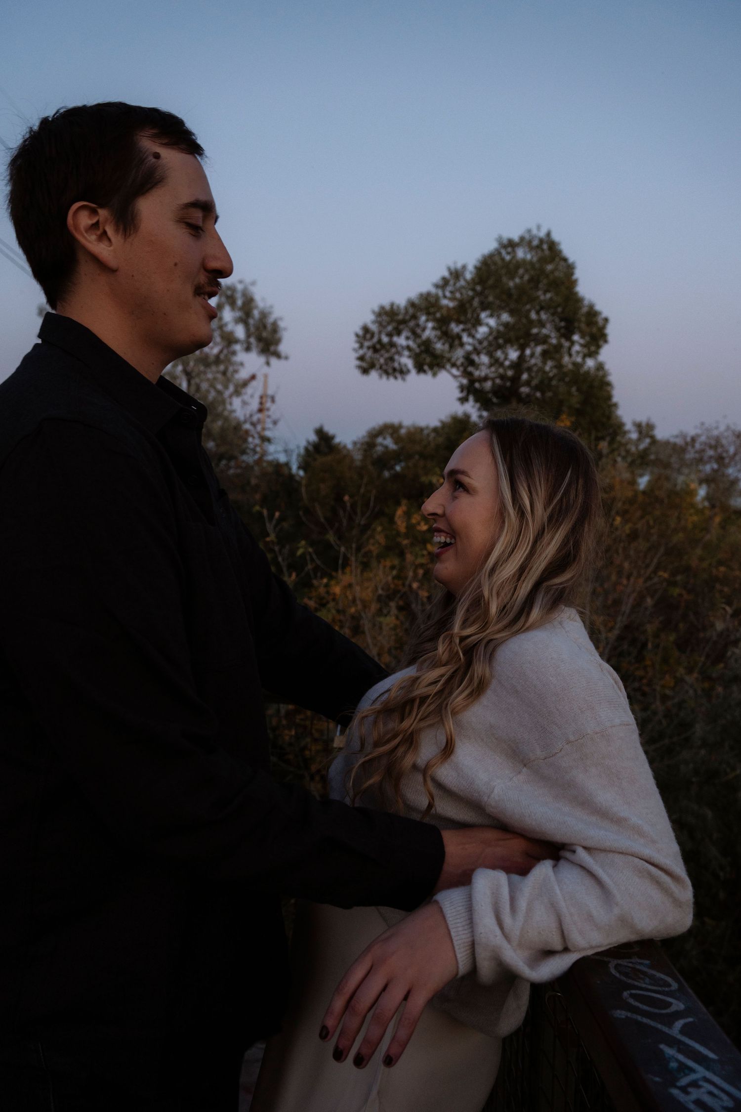 Couple embracing at dusk outdoors, woman in white sweater smiling up at man in black shirt with trees behind.