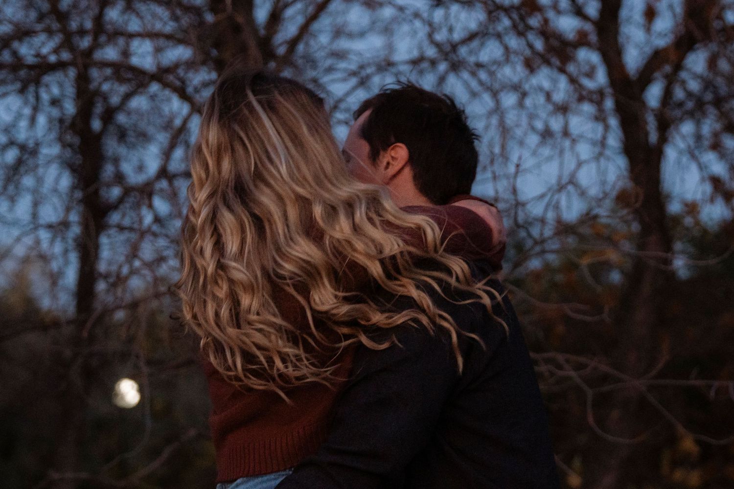Couple embracing outdoors at dusk with bare trees and moonlight in romantic evening setting.