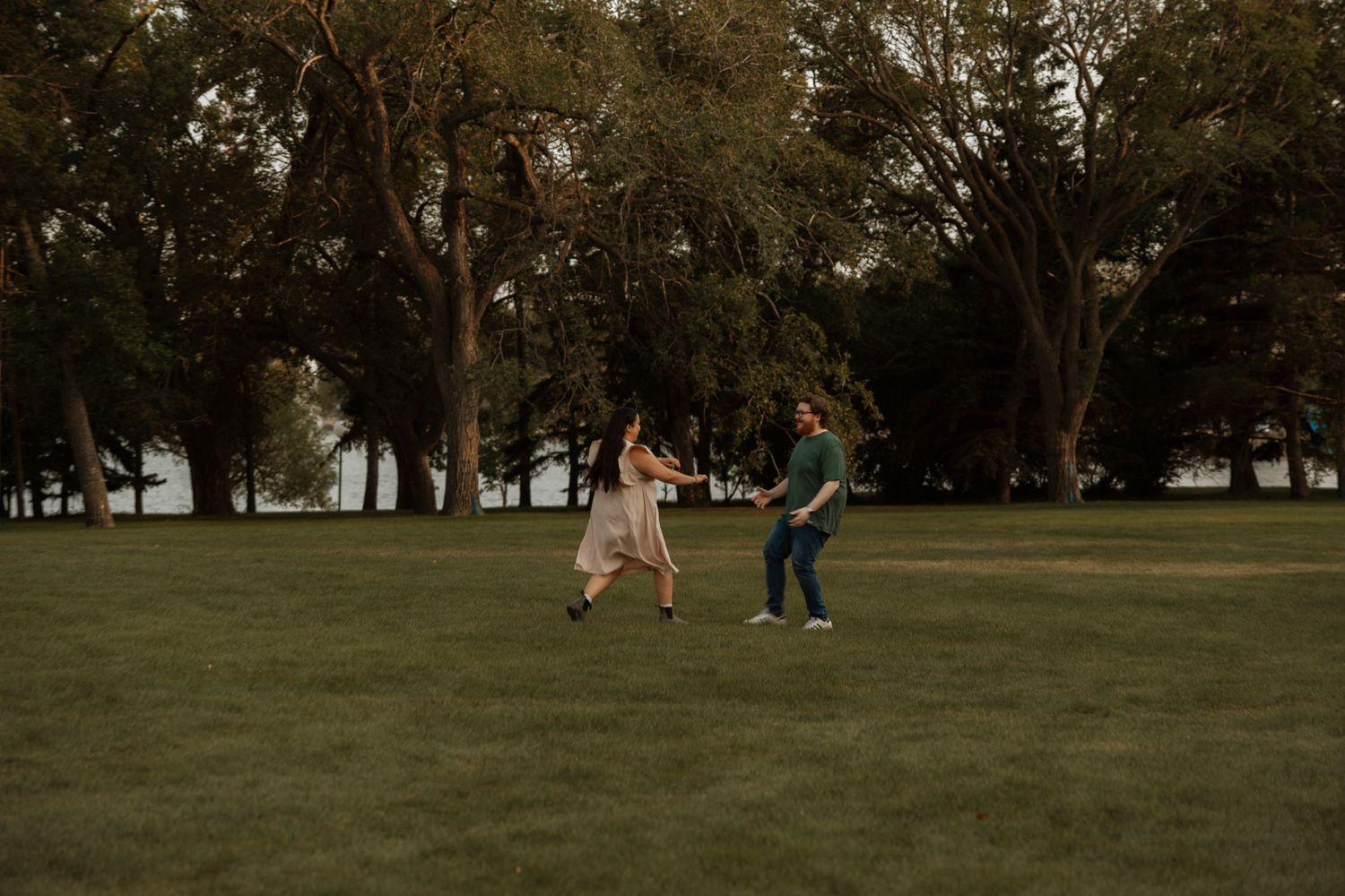 A joyful couple runs hand-in-hand across a lush green park surrounded by tall trees at dusk.