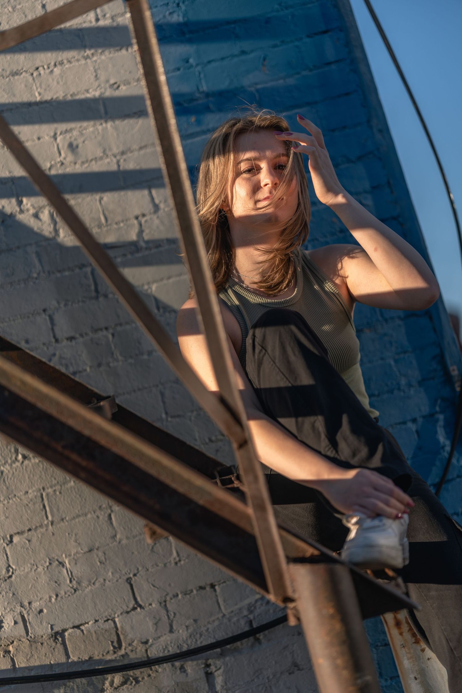 Young woman posing on rustic metal fire escape stairs against blue brick wall in warm golden sunlight.