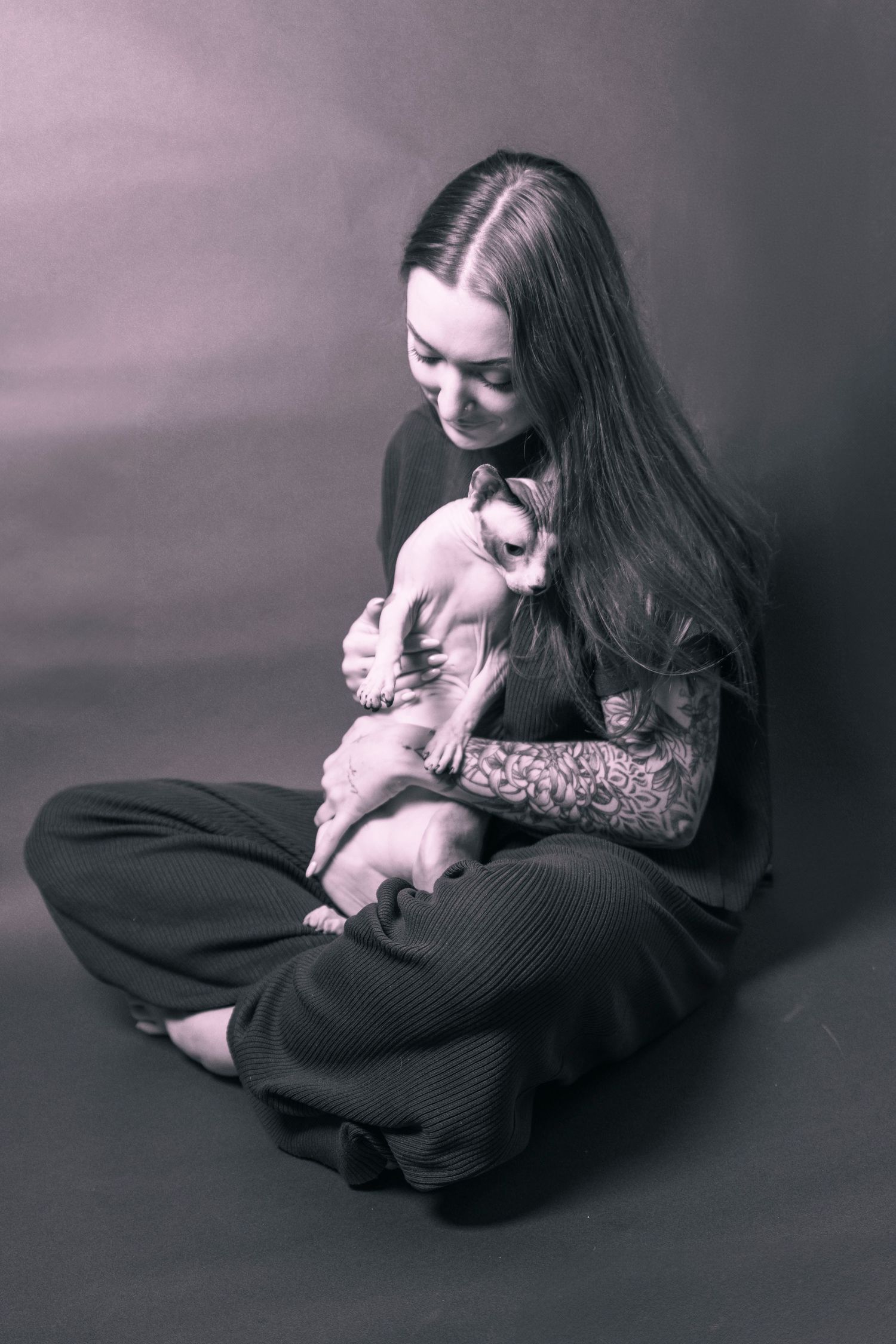 Young woman with long hair and tattoos sits cross-legged holding a hairless Sphynx cat in a moody monochrome studio portrait.