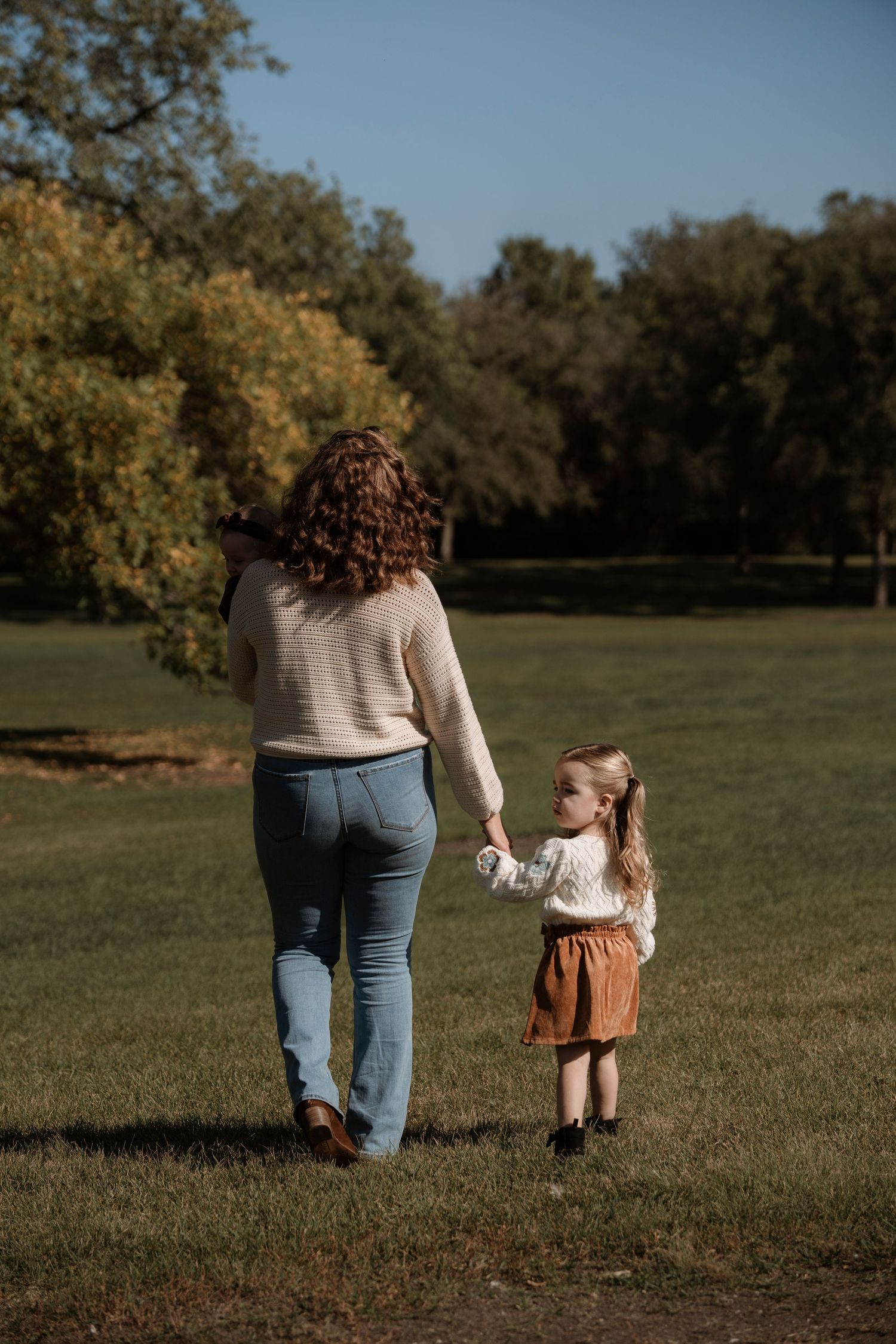A mother and young daughter walk hand-in-hand through a sunny green park, both wearing casual autumn outfits.