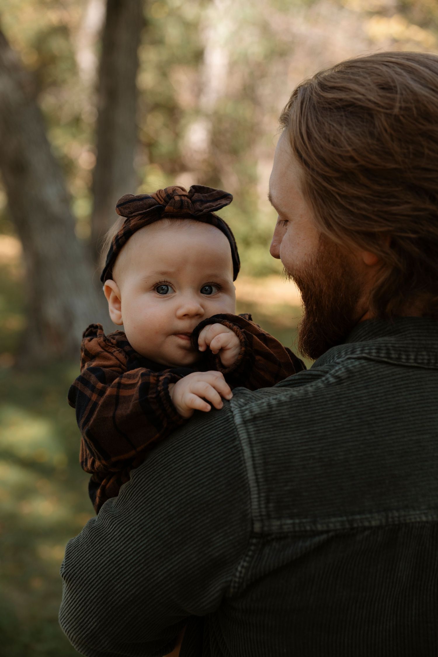 Bearded father holding his baby girl in a dark floral outfit and bow headband outdoors in a forest setting.