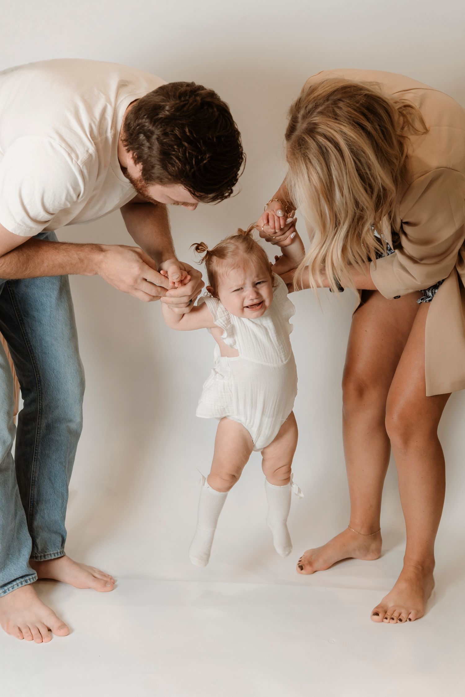 Parents holding baby girl's hands as she stands and smiles in a white studio family photo session.