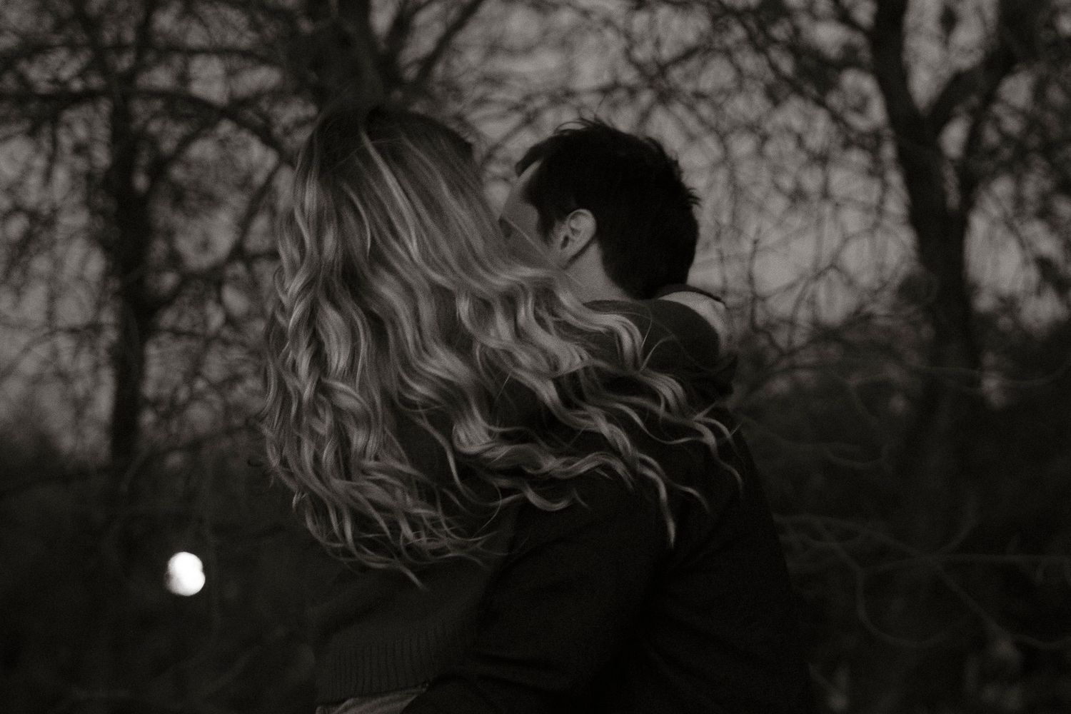 Black and white photo of a couple embracing outdoors at night with bare trees and moonlight in background.