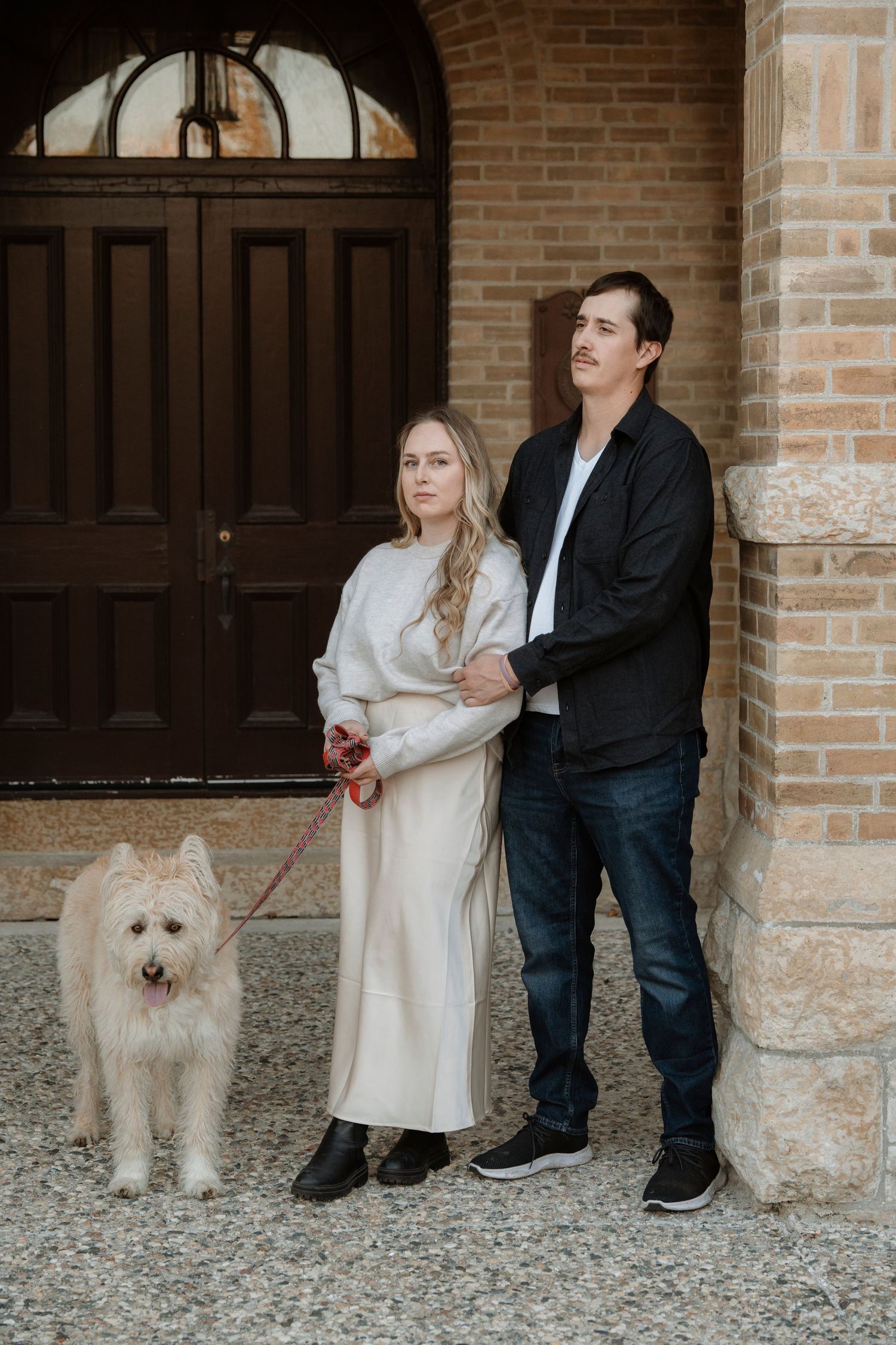Couple with white fluffy dog on leash posing in front of brick building with wooden doors.