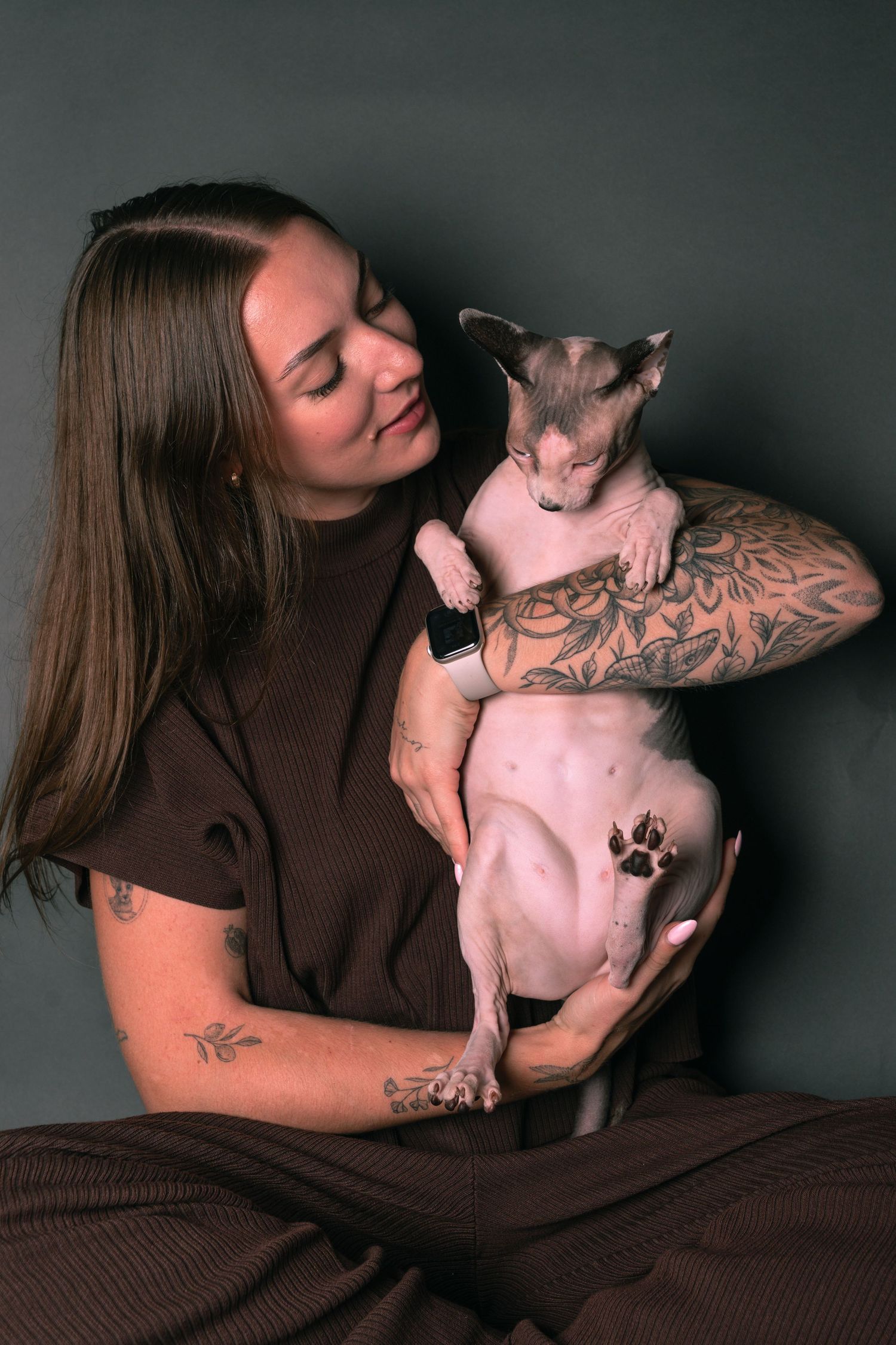 Young woman with tattoos smiling while holding a hairless Sphynx cat in a brown outfit against gray background.