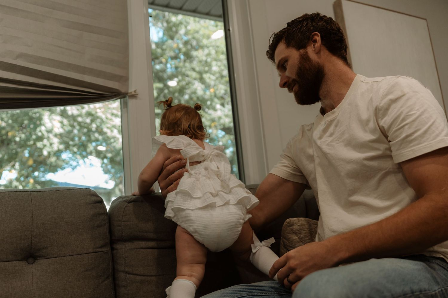Bearded father in white t-shirt interacting with baby in white dress and diaper on gray couch near window.