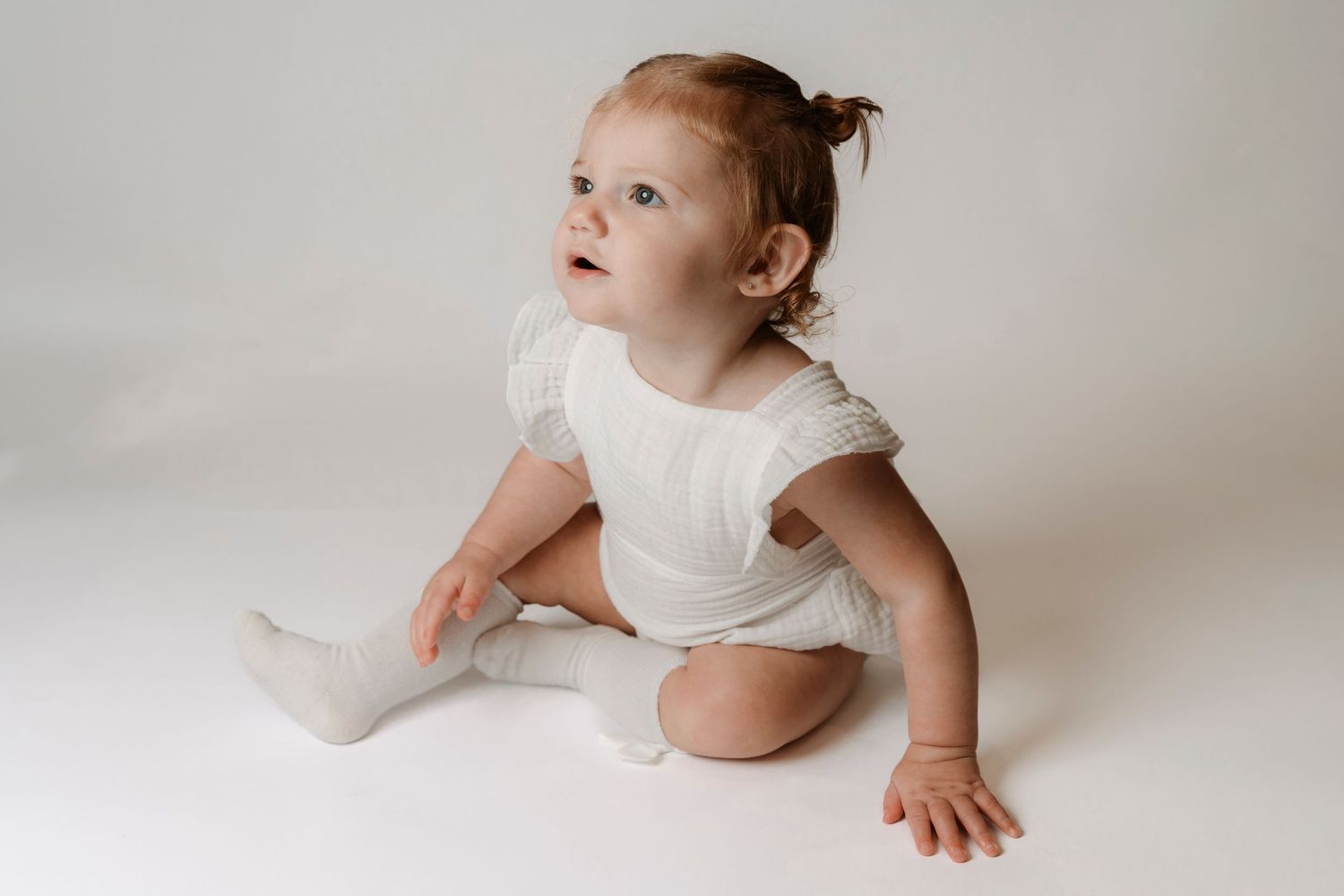 A cute baby girl with red hair sits on a white floor wearing a white romper and socks, looking up curiously.