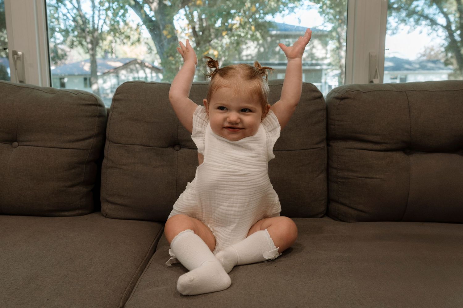 Happy baby girl in white outfit and knee socks sitting on gray sofa with arms raised in celebration.