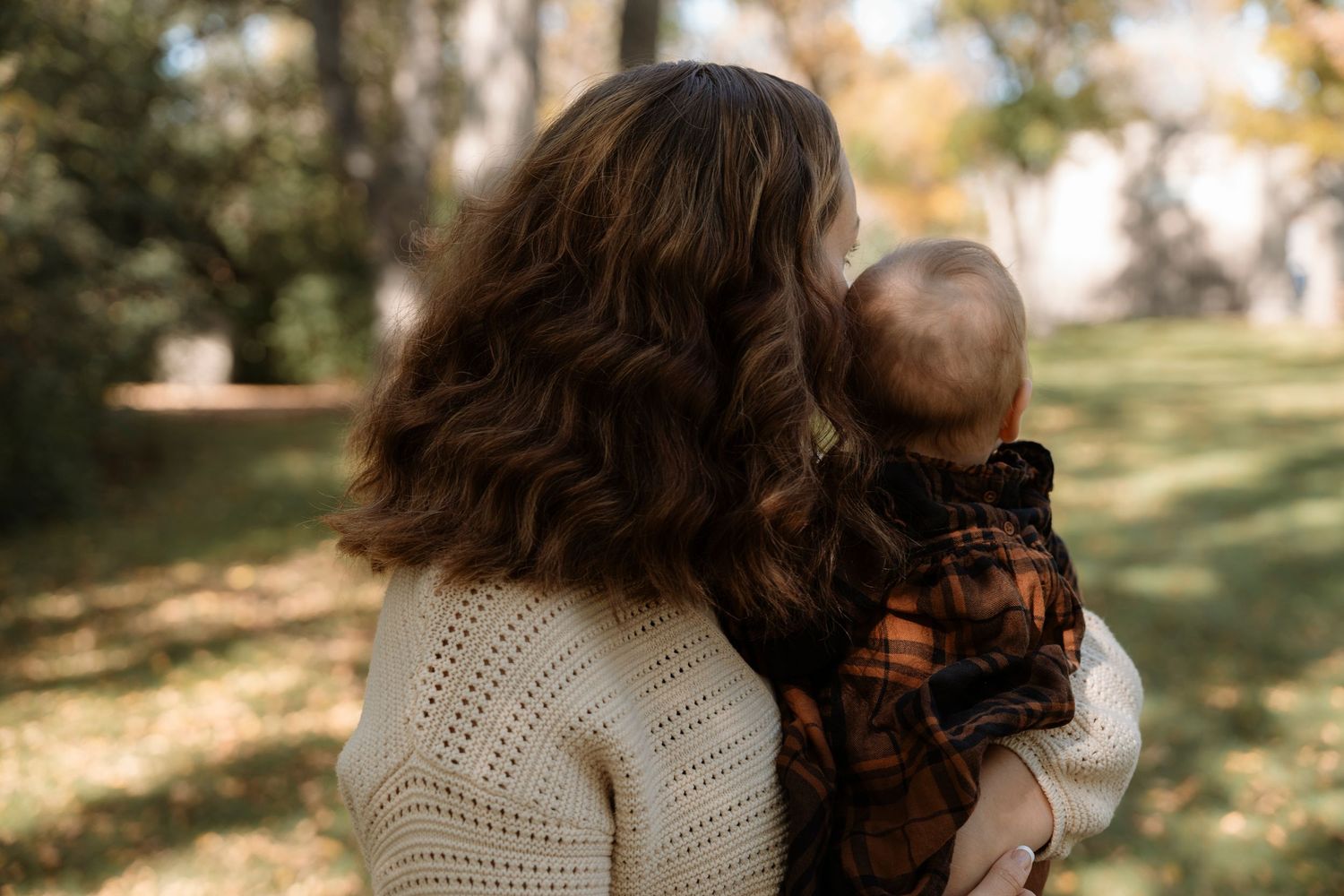 A mother with curly brown hair holds her baby in a park, both facing away in autumn light.