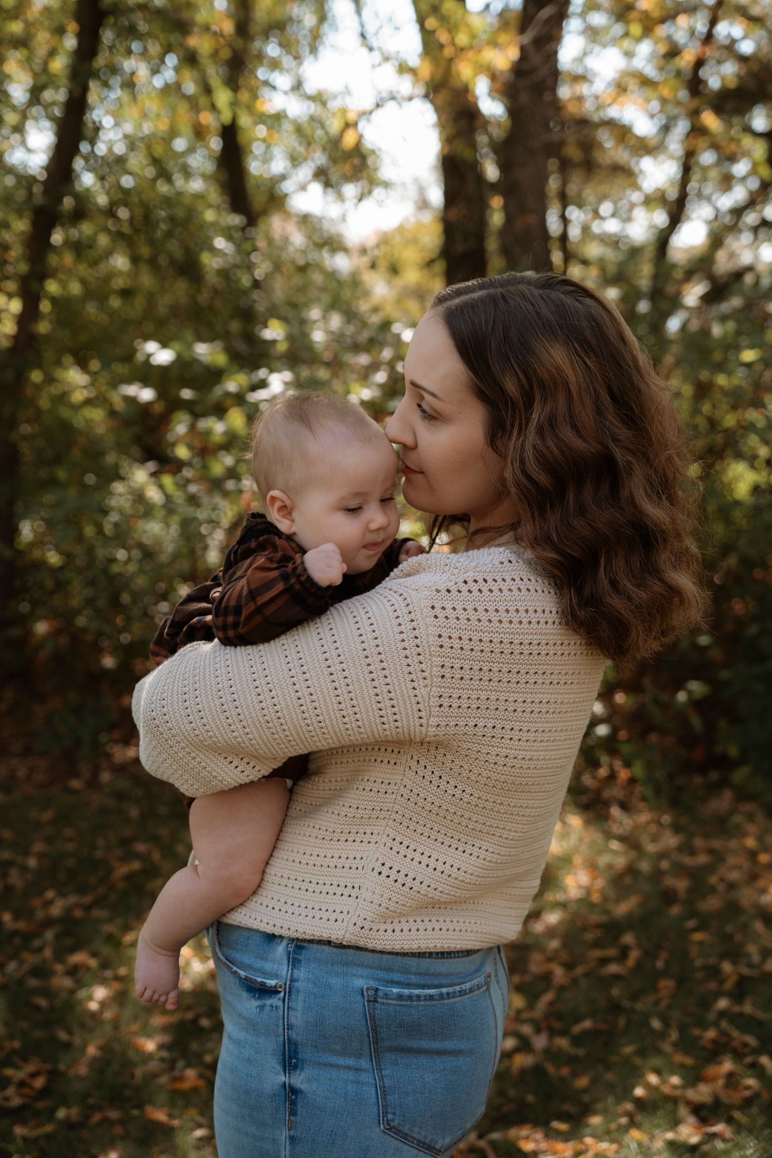 A young mother in a cream knit sweater holds her baby in an autumn forest setting.