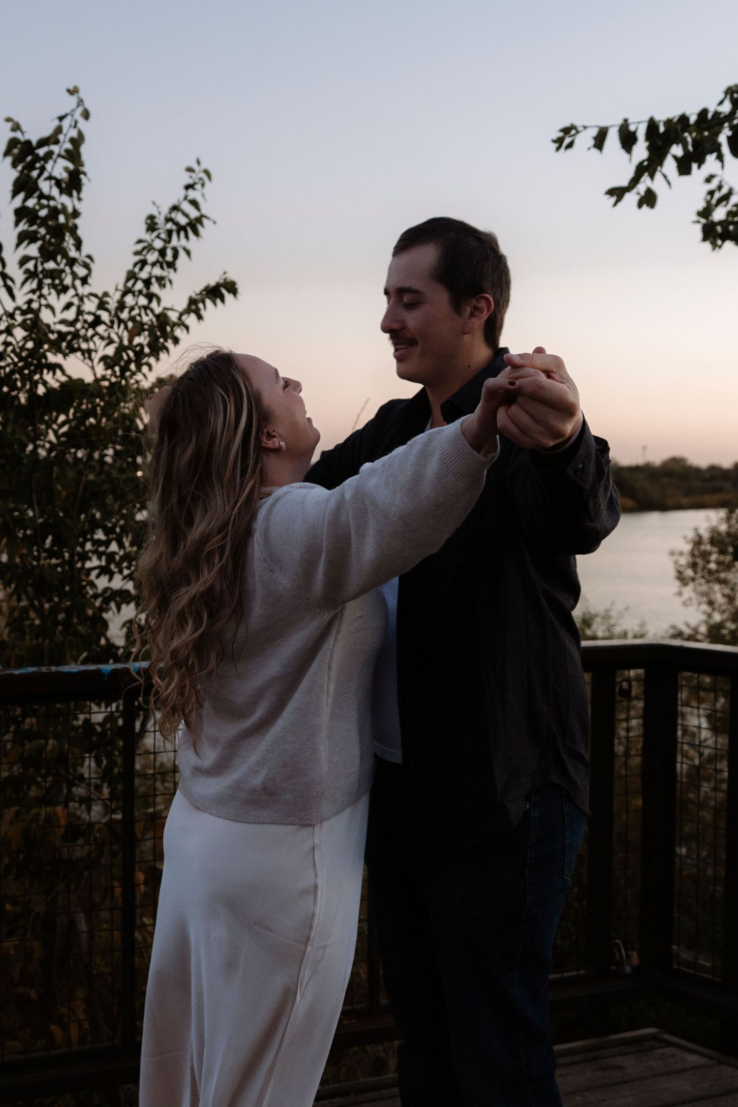 Couple dancing together on a wooden deck at sunset with a lake and trees in the background.