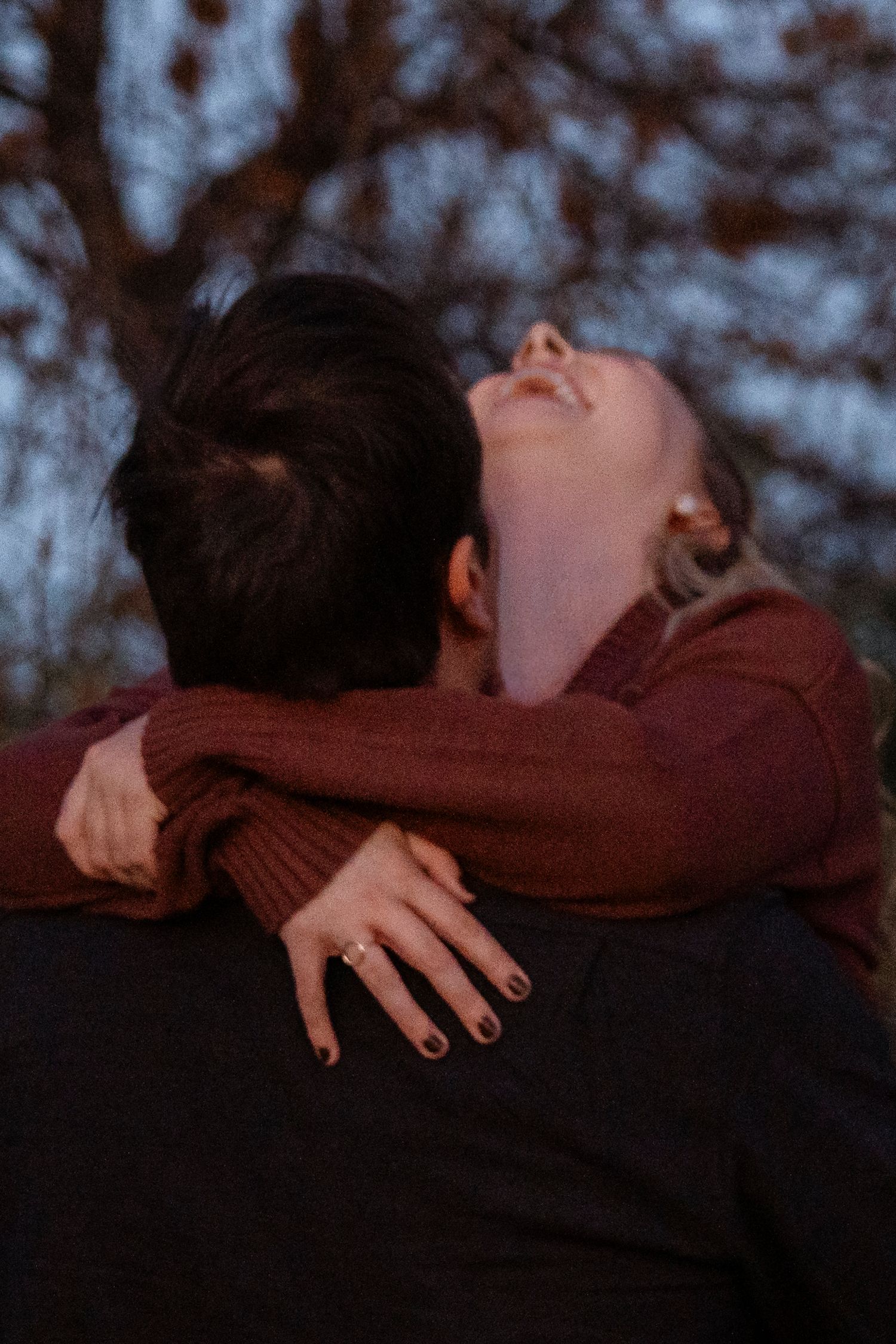 A couple embraces romantically outdoors at dusk, the woman laughing with head tilted back, wearing a burgundy sweater and ring.