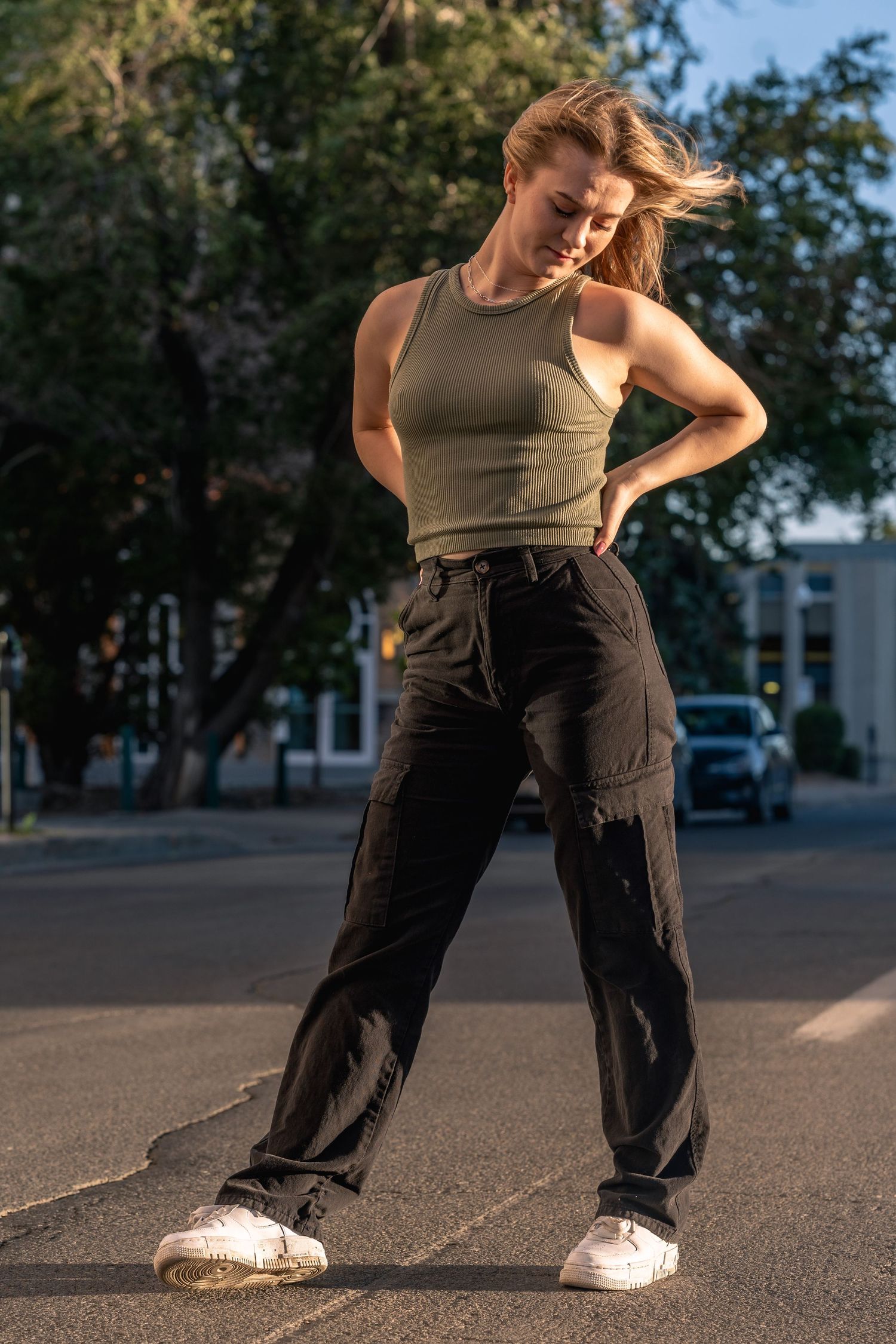 Young woman in olive tank top and black cargo pants dancing outdoors on a street at golden hour.
