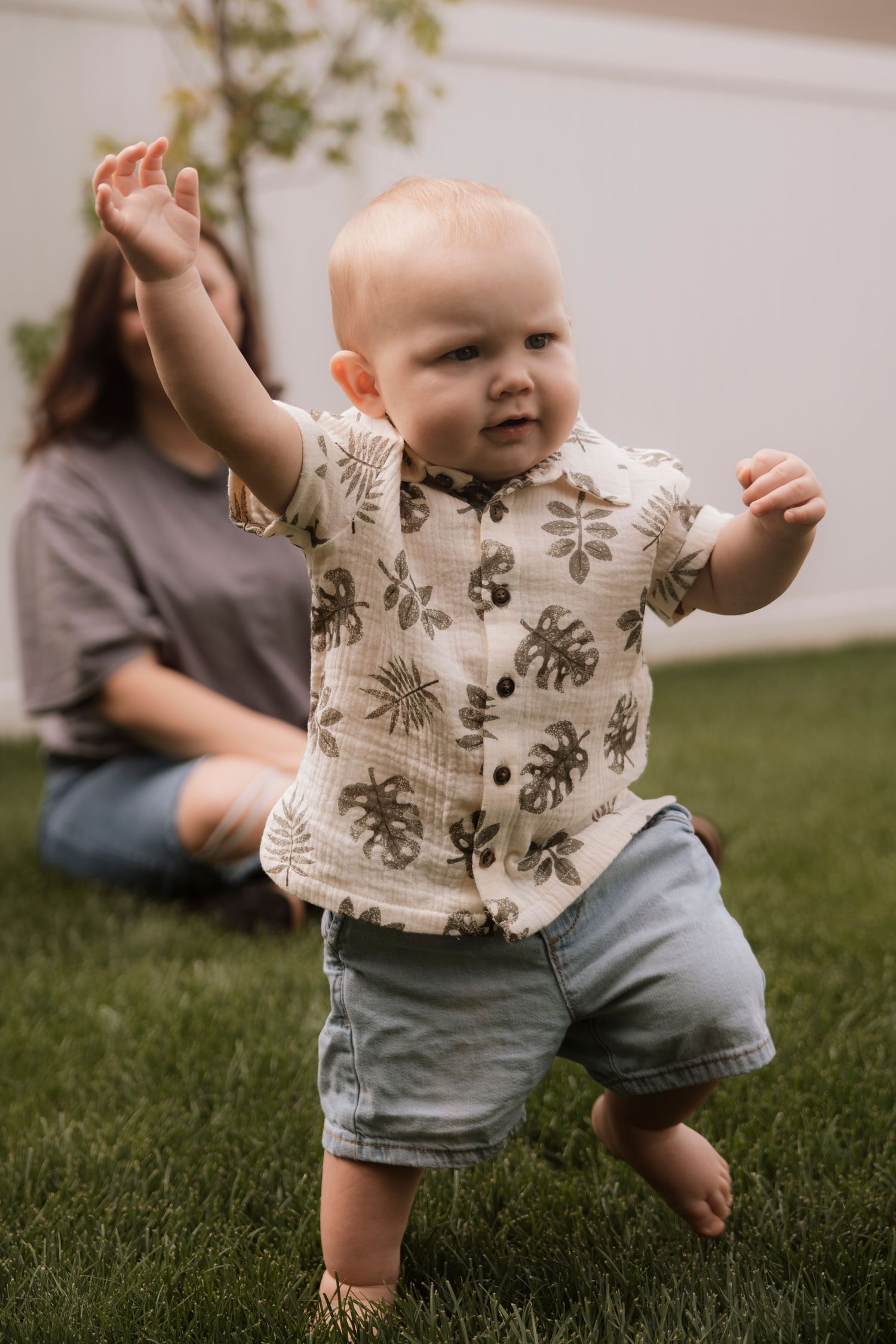 Baby in floral shirt and denim shorts explores while crawling on green grass. Regina Saskatchewan Family portrait photographer.