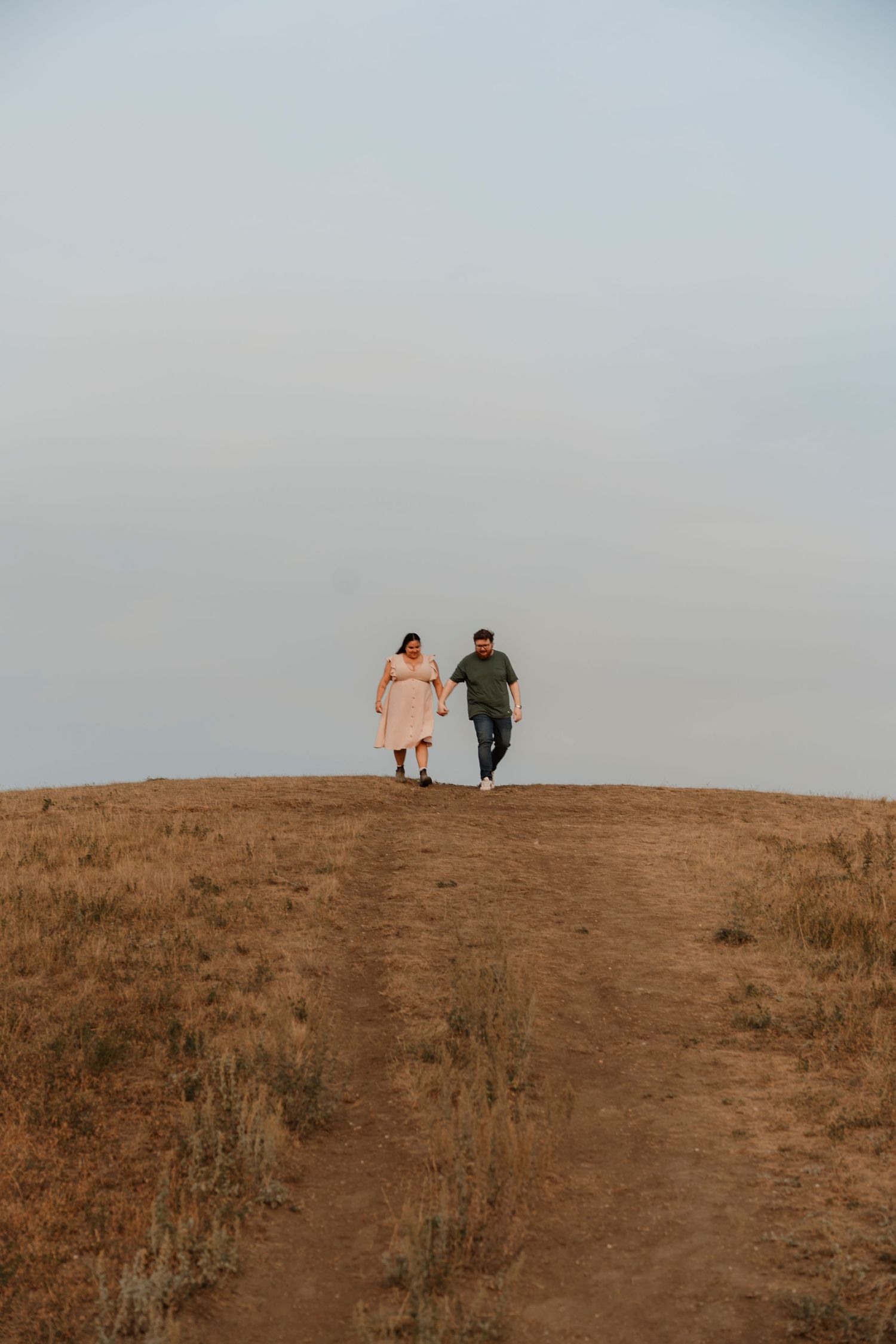 Couple holding hands walking on a dirt path through golden fields under a soft gray sky.