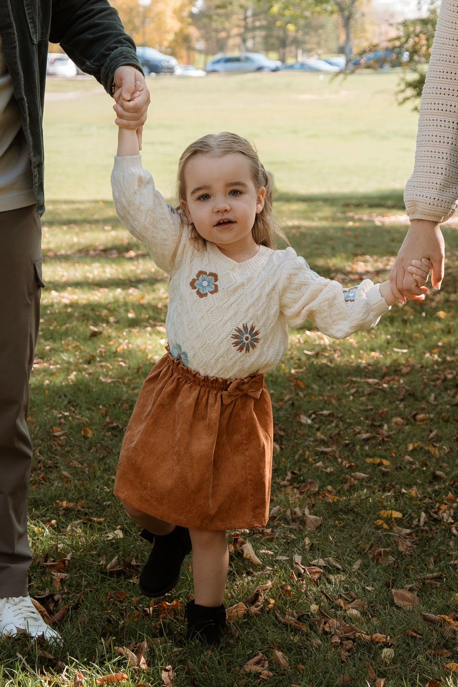 Toddler girl in floral sweater and brown skirt holds hands with parents while walking in autumn park.