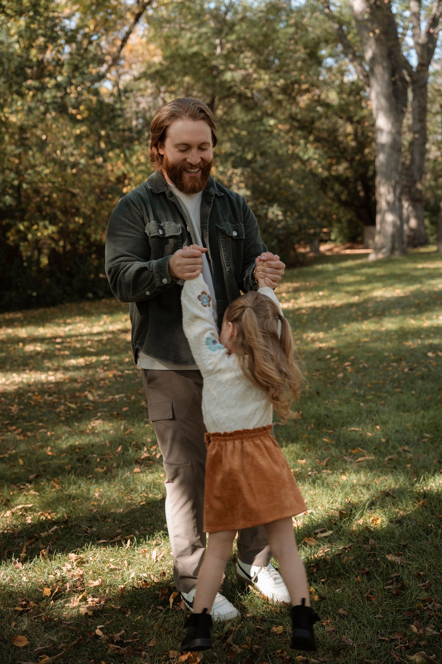 A bearded man smiles while dancing with a young girl in a sunny autumn park, both wearing casual fall outfits.