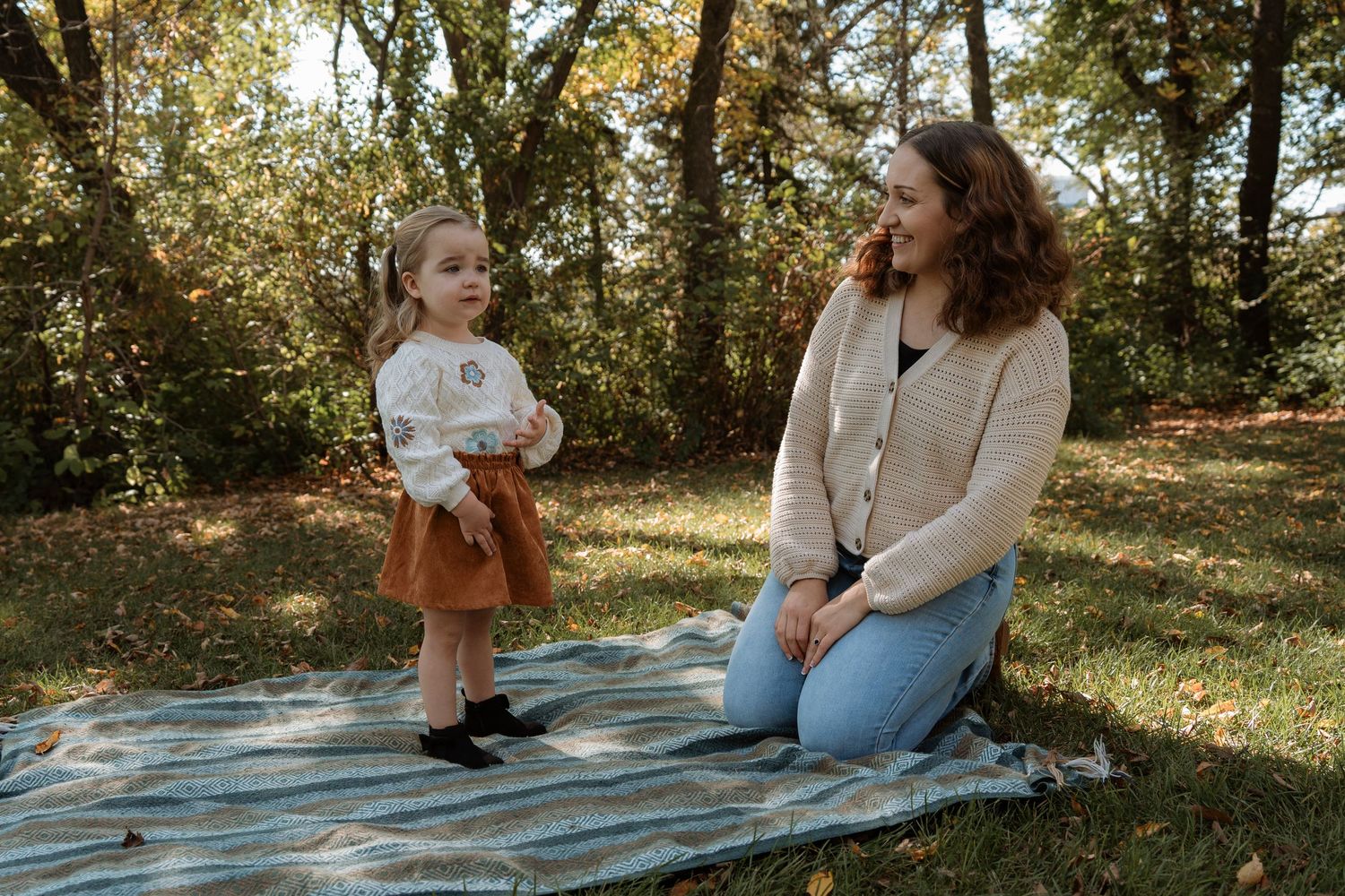 A woman kneels on a blanket outdoors while a young toddler girl in a rust skirt stands nearby in a park setting.
