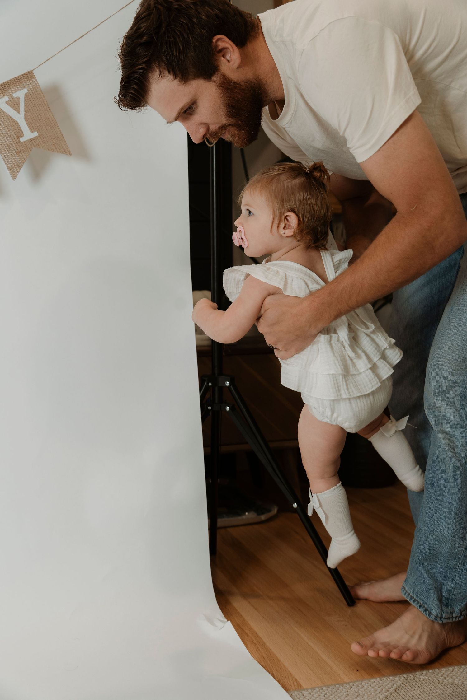 Father helping baby girl take first steps near a door, both barefoot indoors on hardwood floor.