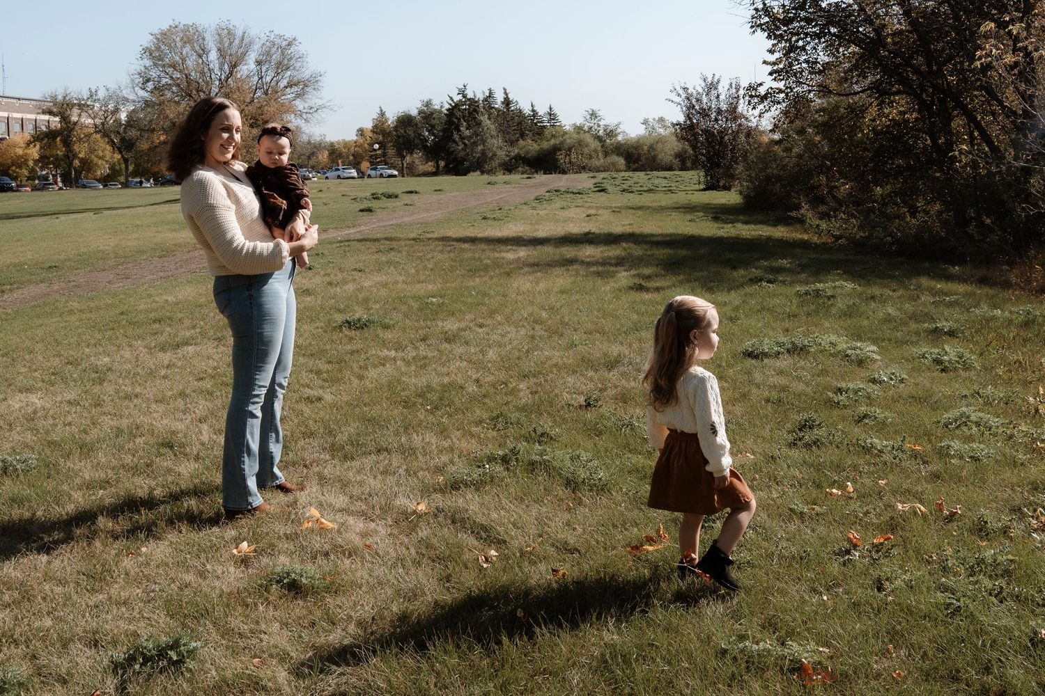 A mother holding a baby while a young girl runs toward her in a green park on a sunny autumn day.