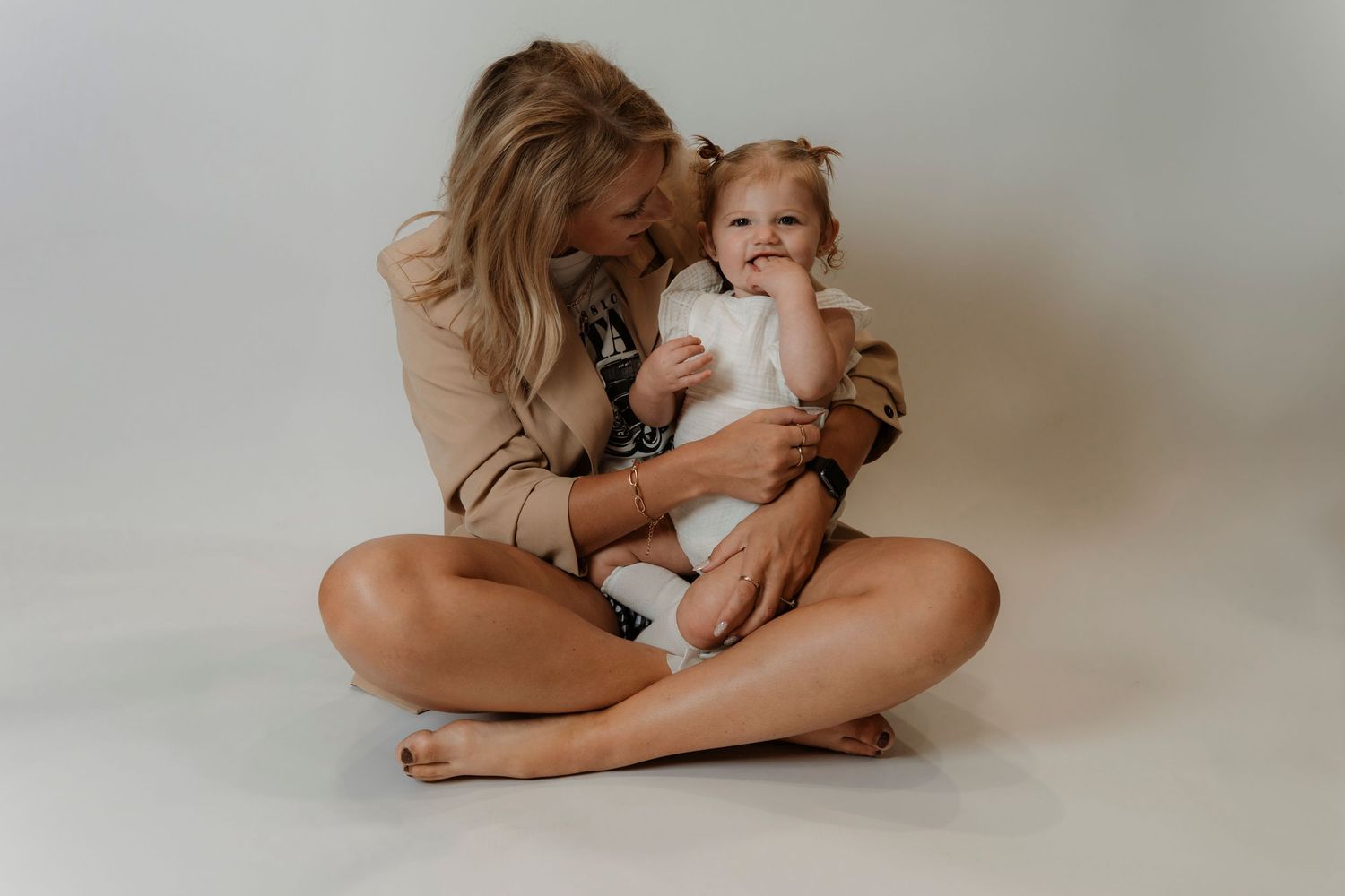 A woman sits cross-legged on a white background, kissing a baby girl in a white outfit on the cheek.