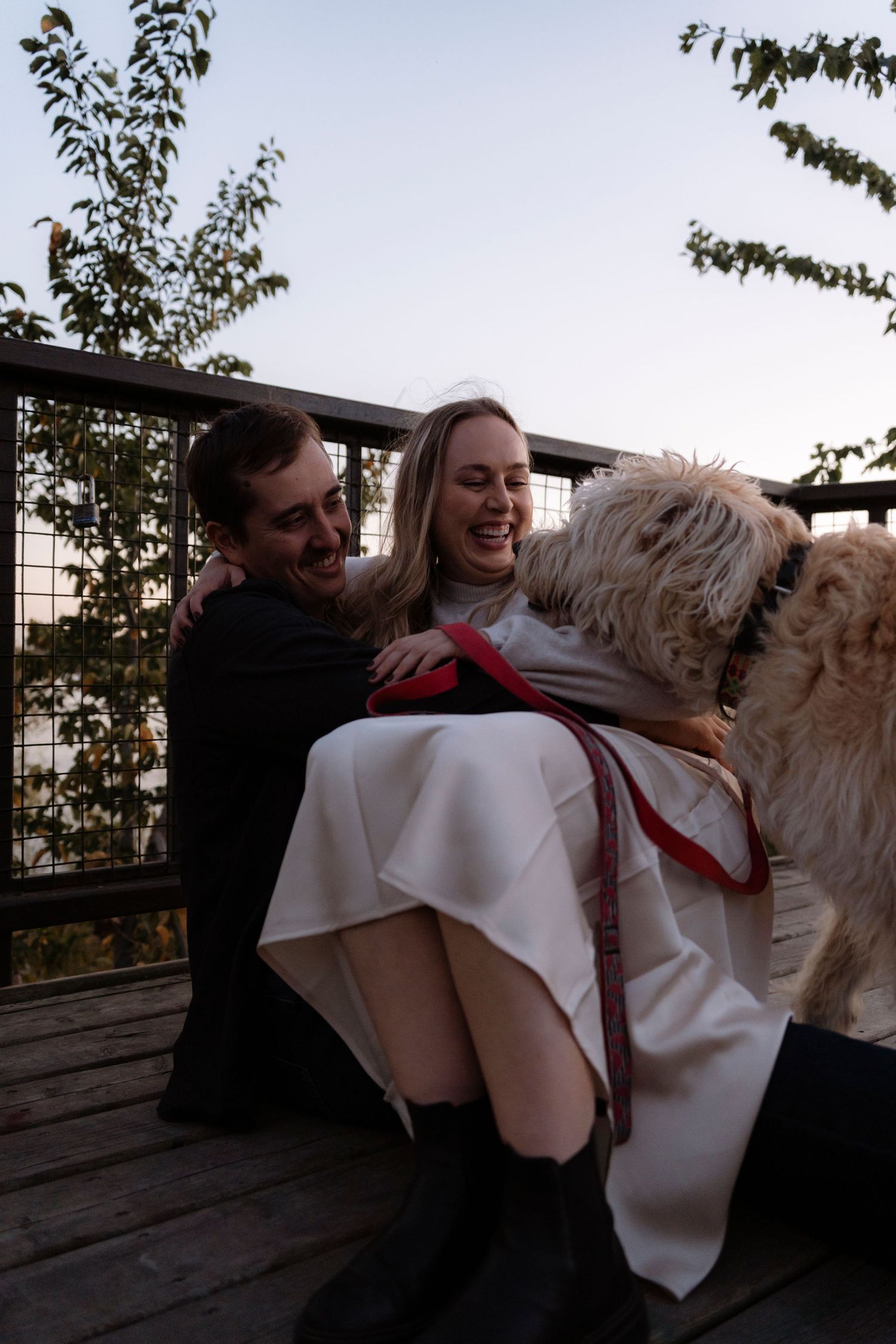 A laughing couple on a wooden deck with a fluffy dog on a red leash during a romantic outdoor moment.
