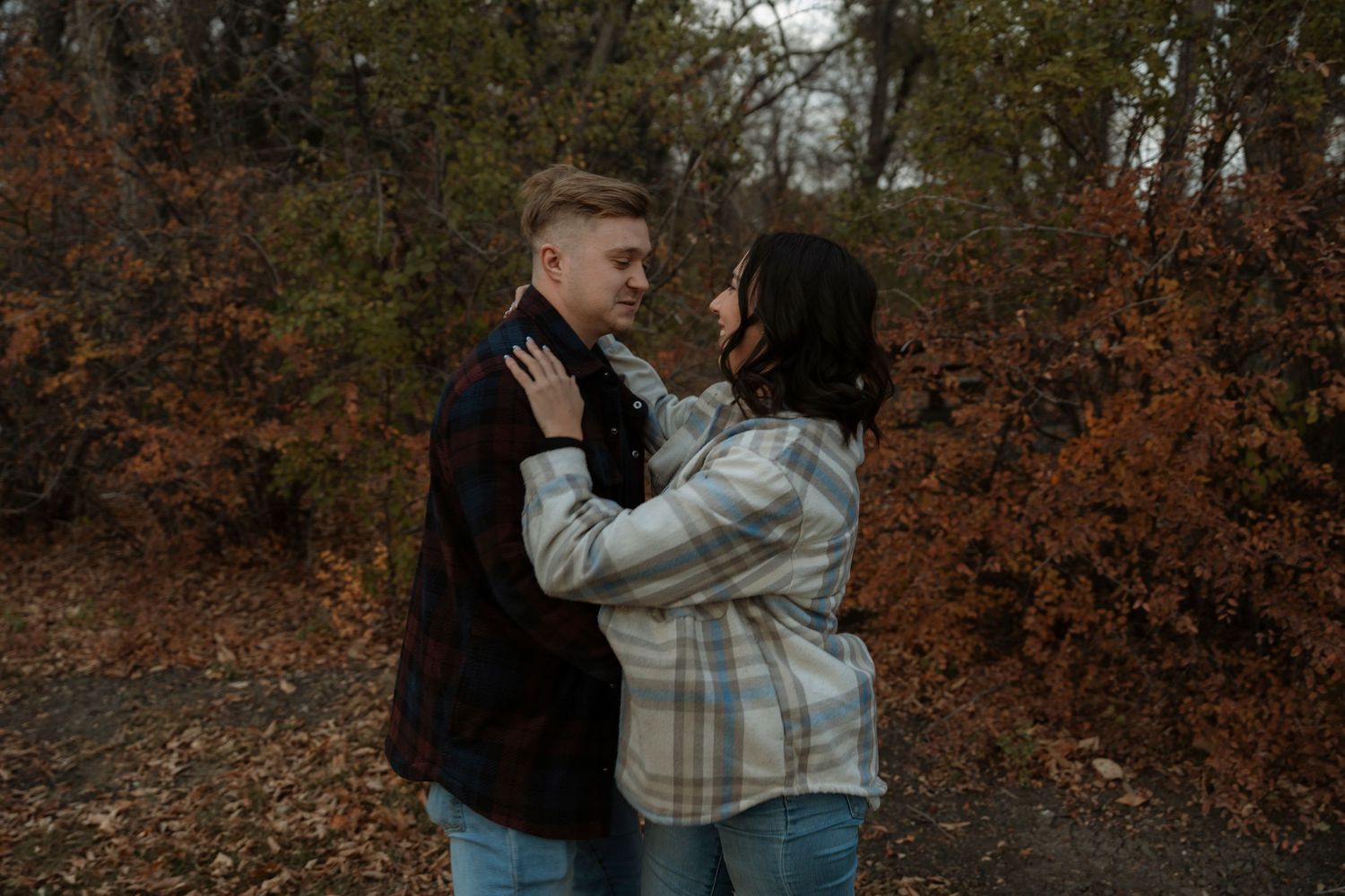 A couple embraces romantically outdoors among autumn foliage, gazing into each other's eyes on a cloudy fall day.