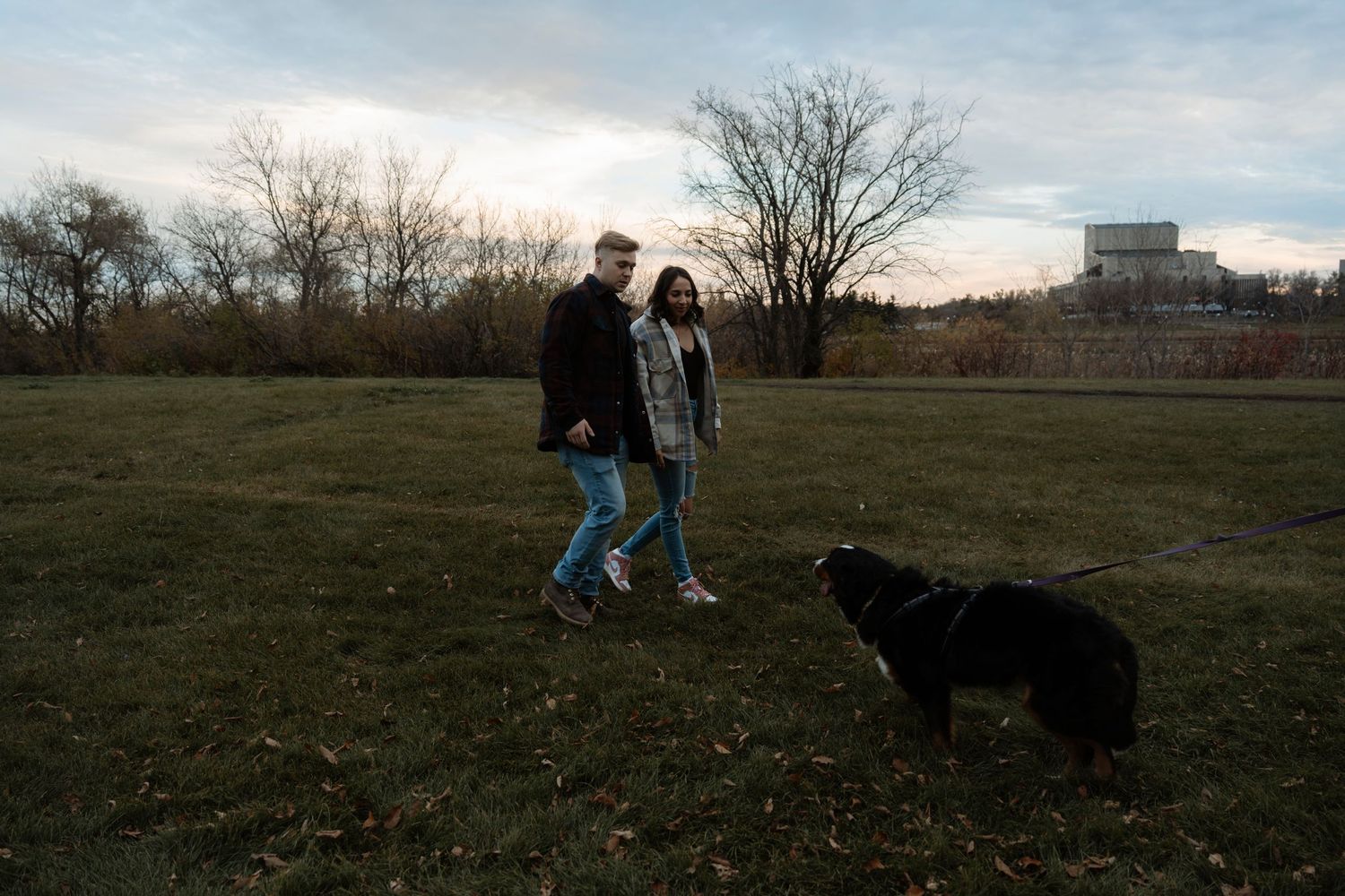 Couple walking black dog in park at dusk, bare trees and cloudy sky in background.