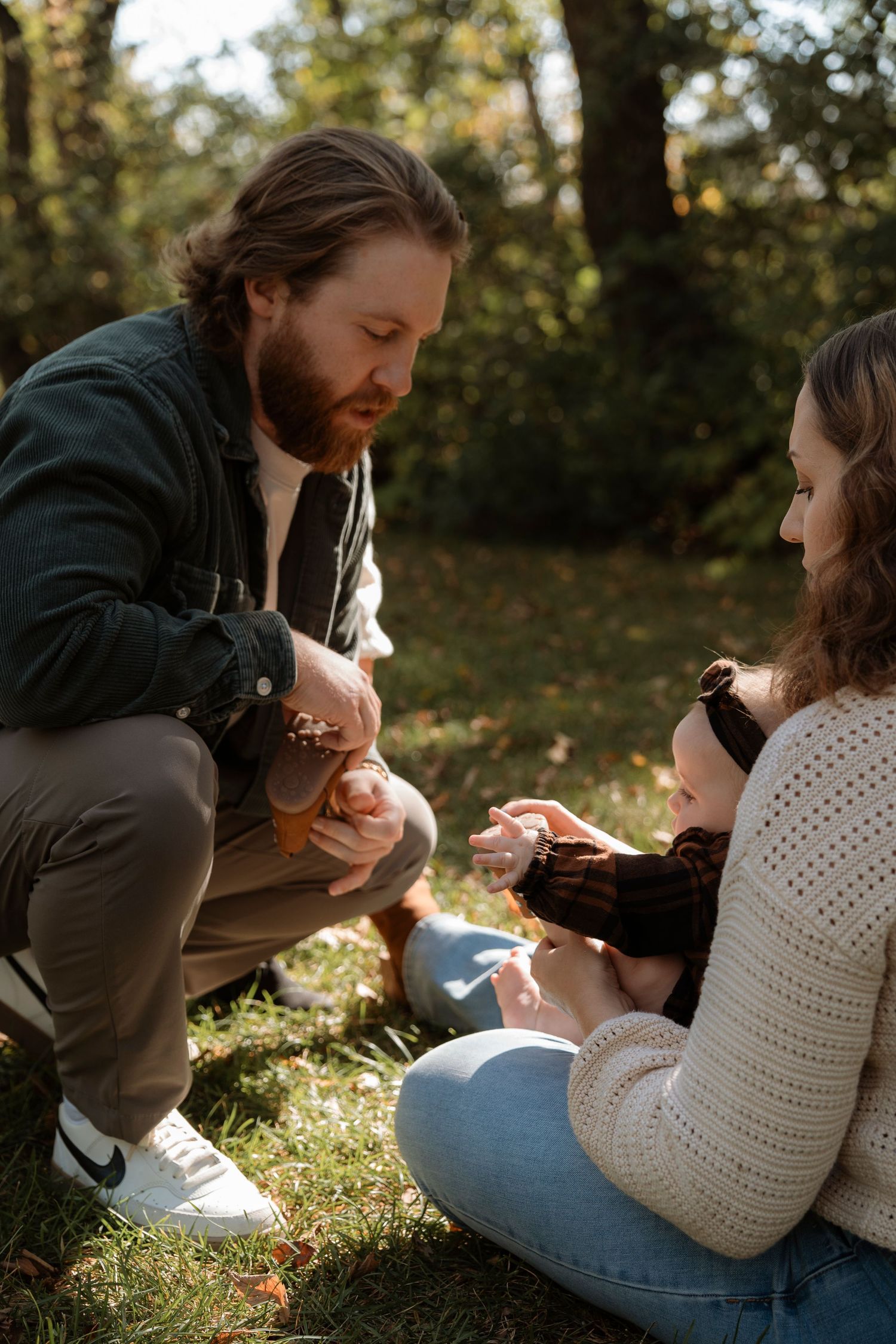 A bearded father and mother sit outdoors on grass, lovingly interacting with their baby in a sunny autumn park.
