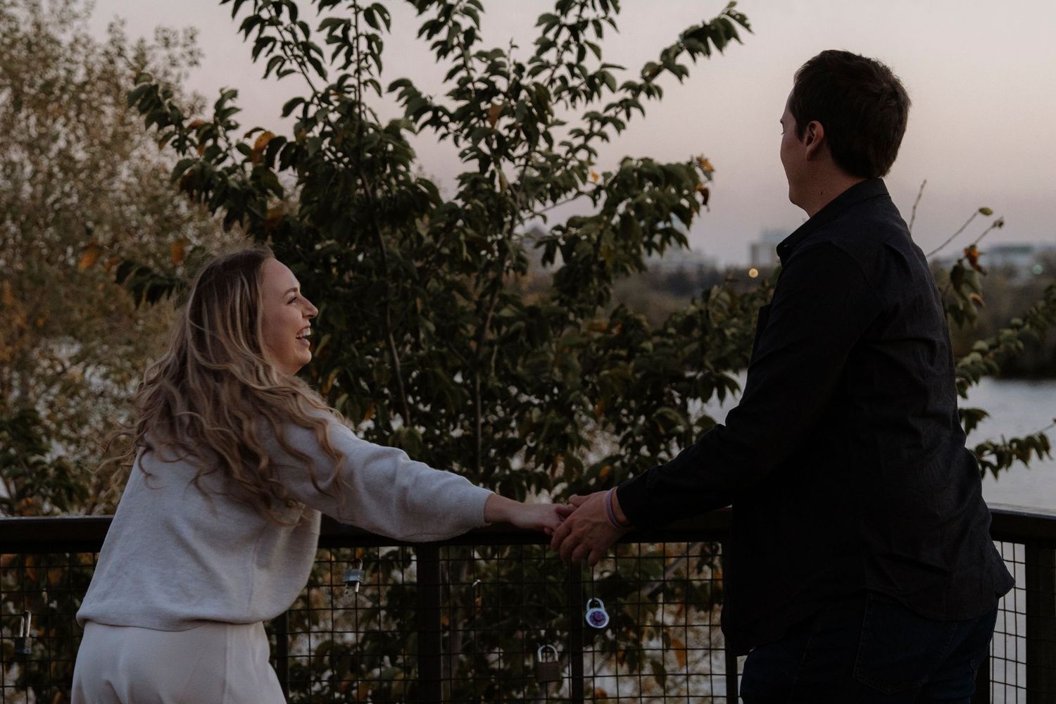 Couple holding hands on an outdoor balcony at sunset, surrounded by autumn trees and a scenic waterfront view.