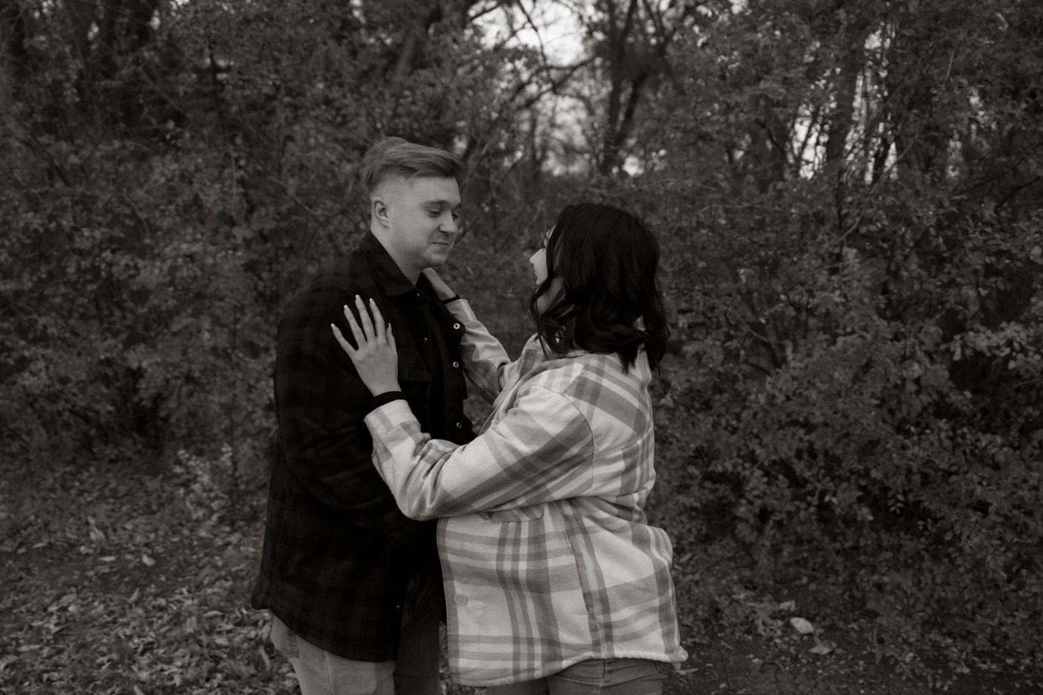 Black and white photo of a couple sharing a tender moment in a wooded outdoor setting.