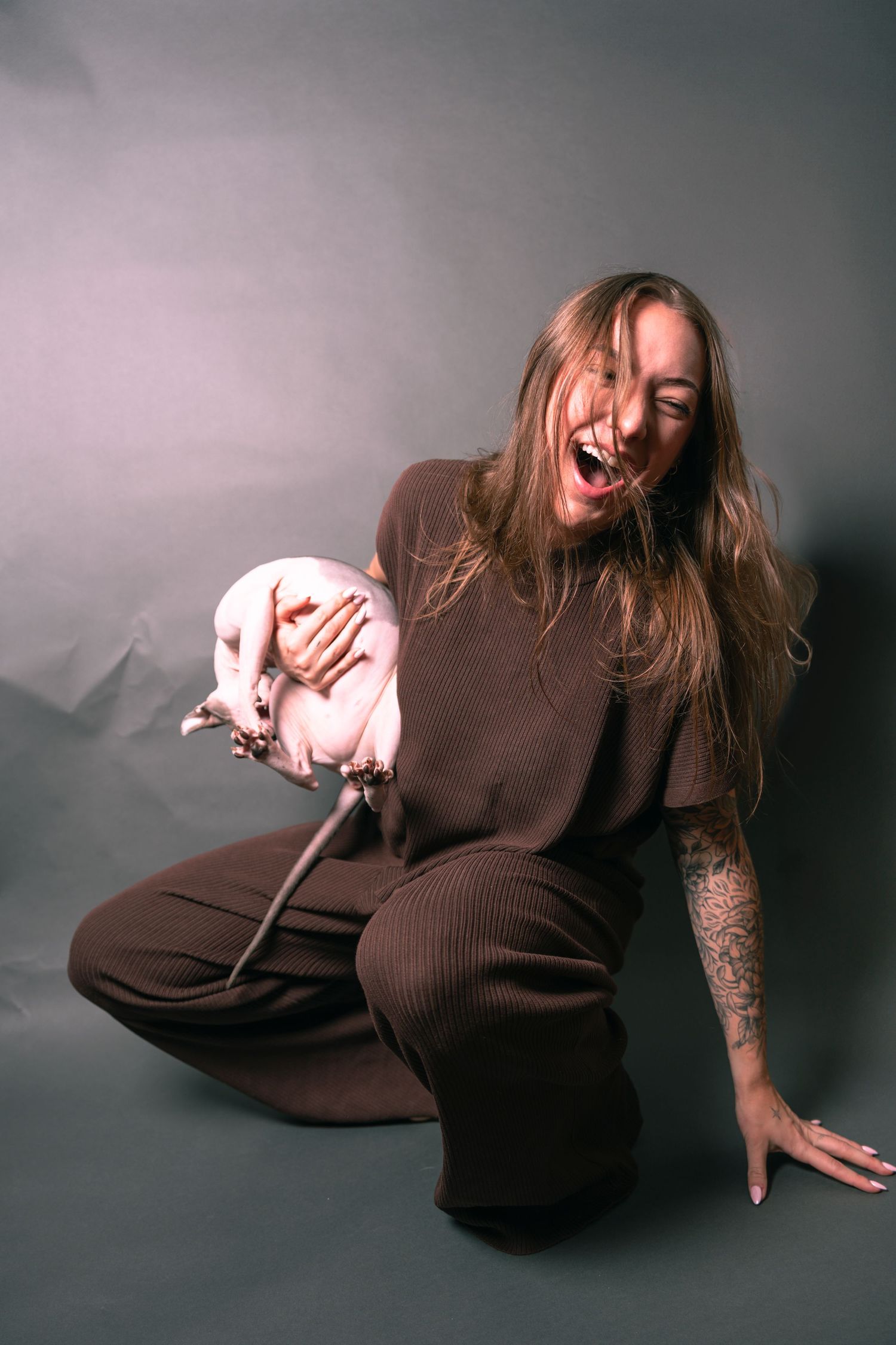 Woman in brown outfit laughing while holding a hairless cat, sitting on floor with tattoos visible.