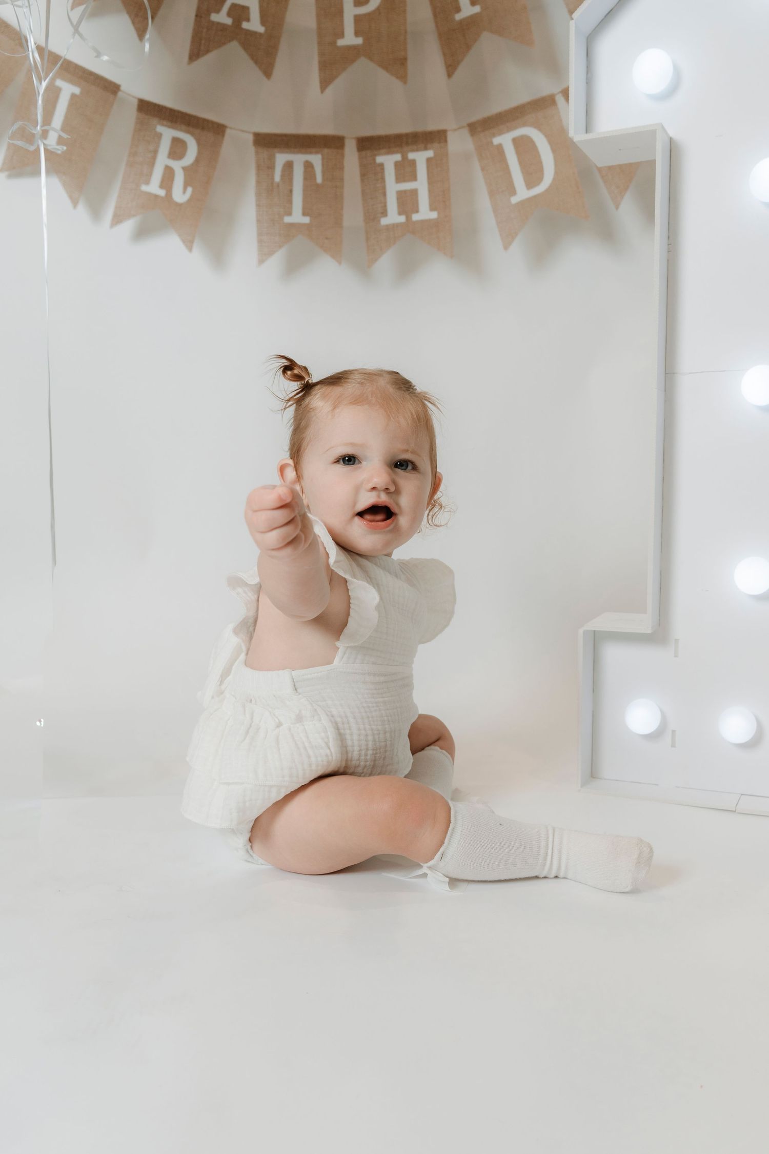 Adorable baby girl in white knit outfit sitting at first birthday party with burlap Happy Birthday banner.
