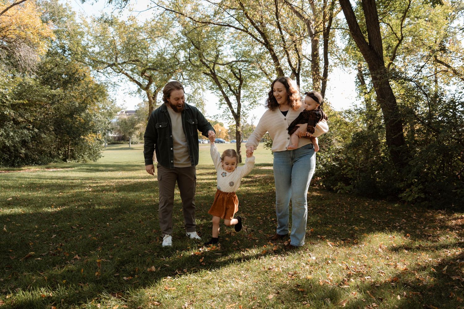 Family of four walking in a sunny autumn park, parents holding toddler's hands while carrying a baby.