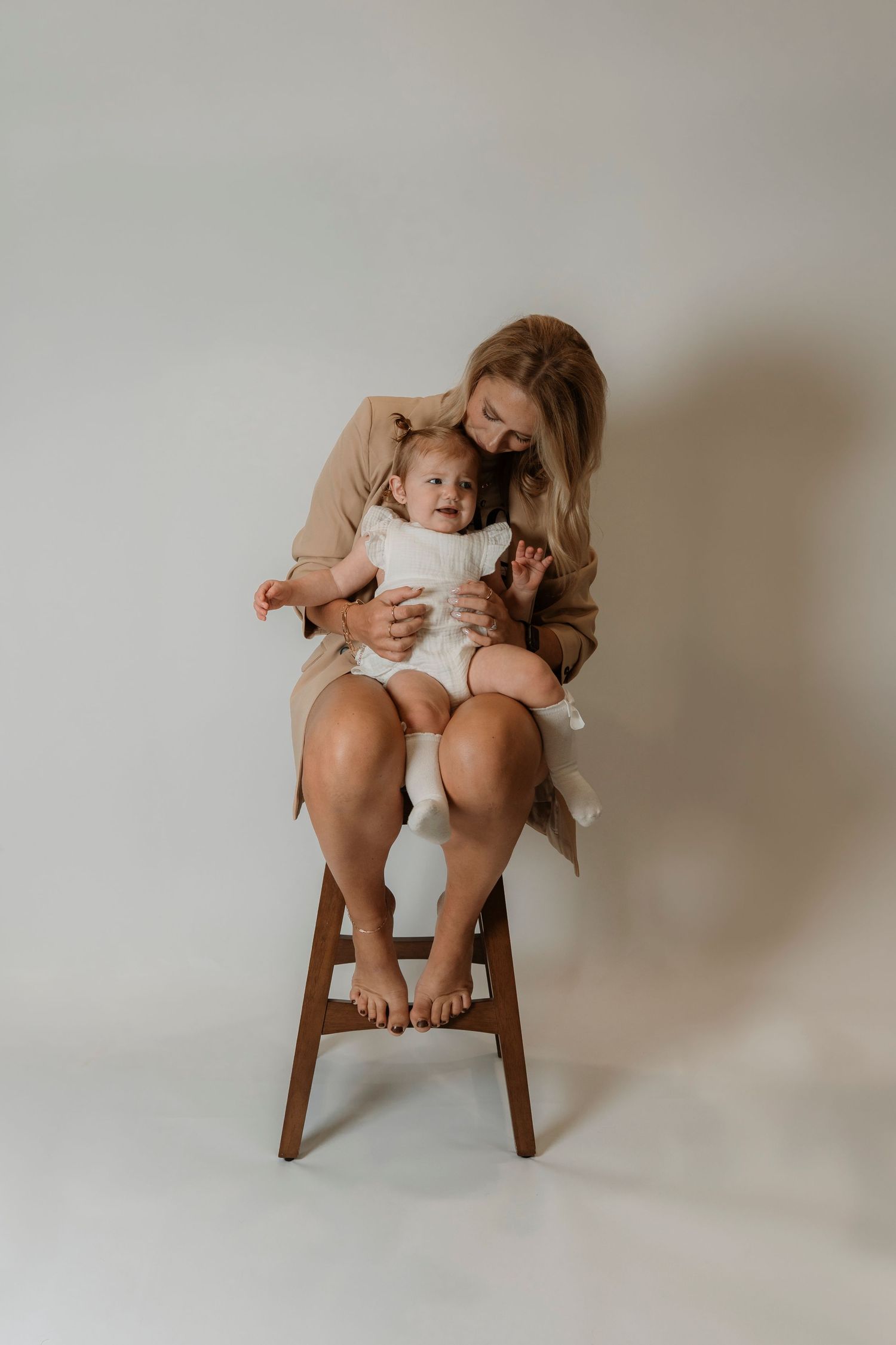 A blonde mother sits on a wooden stool holding her newborn baby dressed in white against a neutral background.