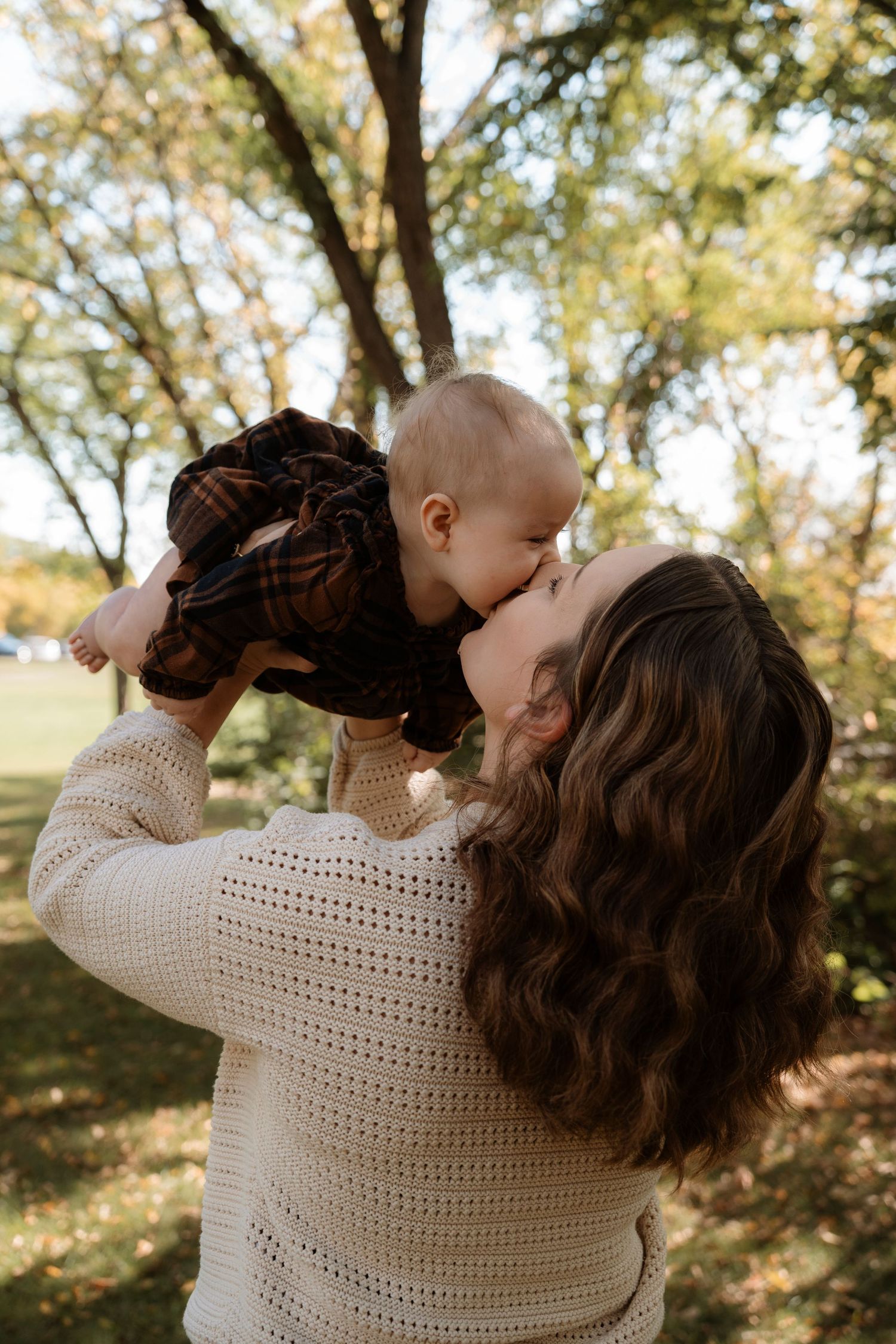 A mother in a cream sweater lifts her baby for a kiss outdoors among autumn trees.