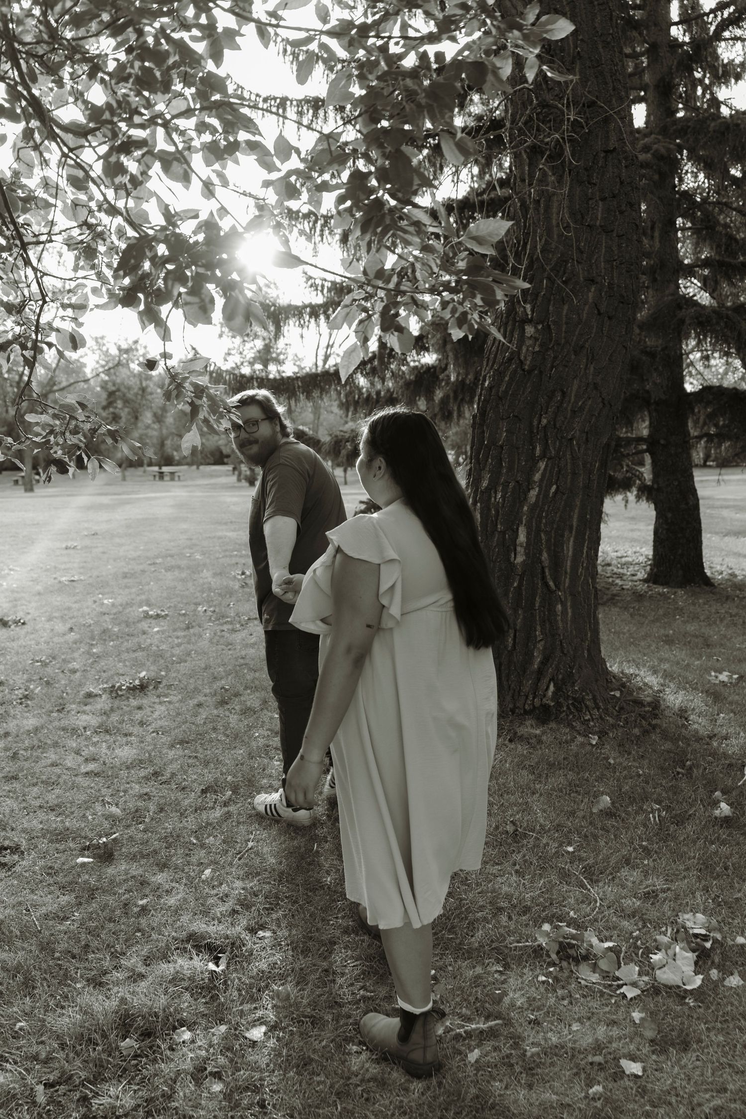 Black and white photo of a couple standing near large trees in a park, woman in dress facing man.