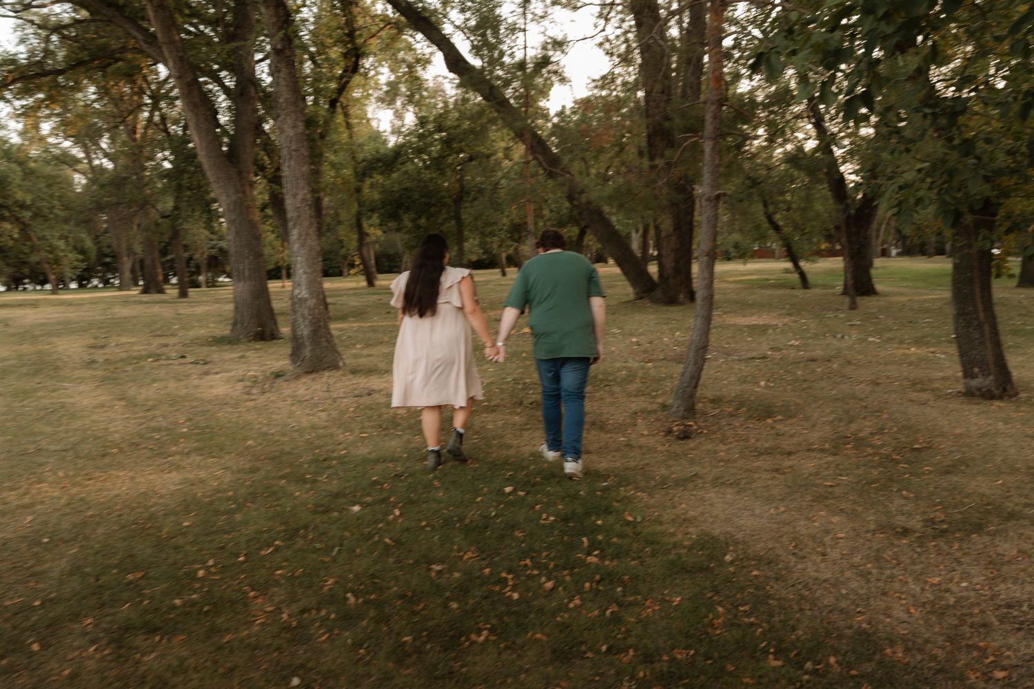 Couple holding hands walking through a lush green park with tall trees on a cloudy day.