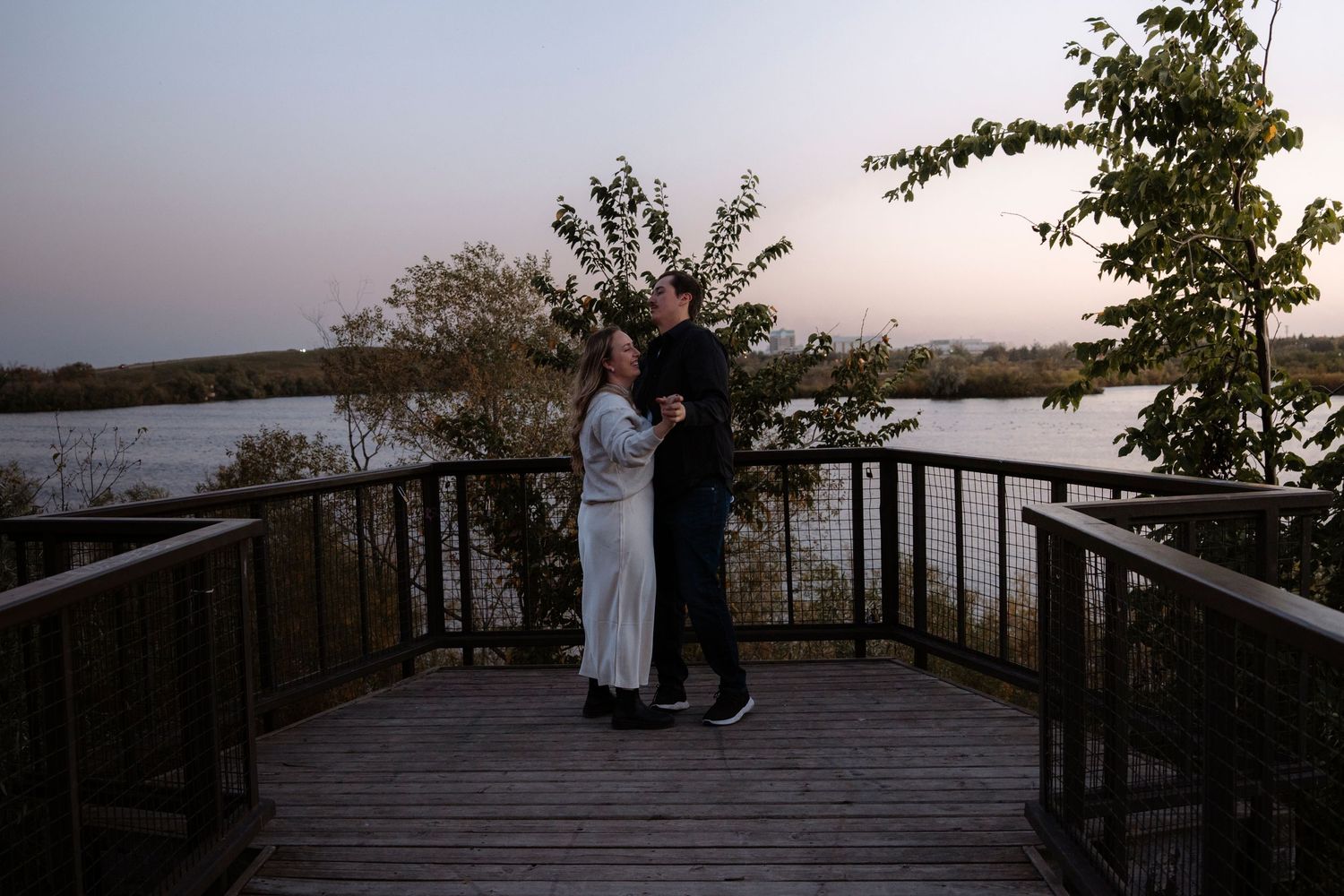 Couple embracing on a wooden deck overlooking a serene lake at dusk with trees in the background.